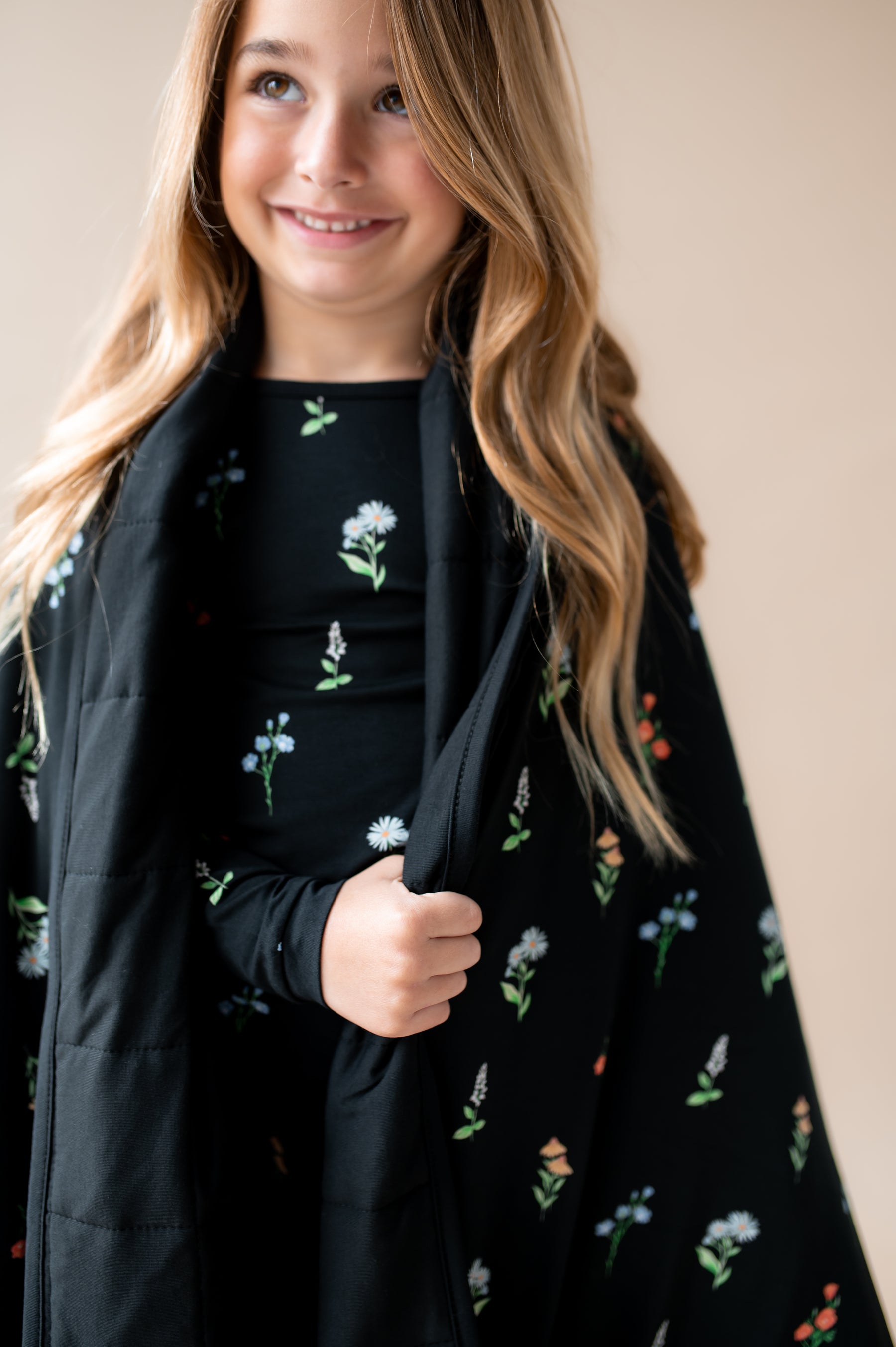 Girl smiling with Youth Blanket in Midnight Wildflower 2.5 print over her shoulders wearing a similar print pajamas on a beige background