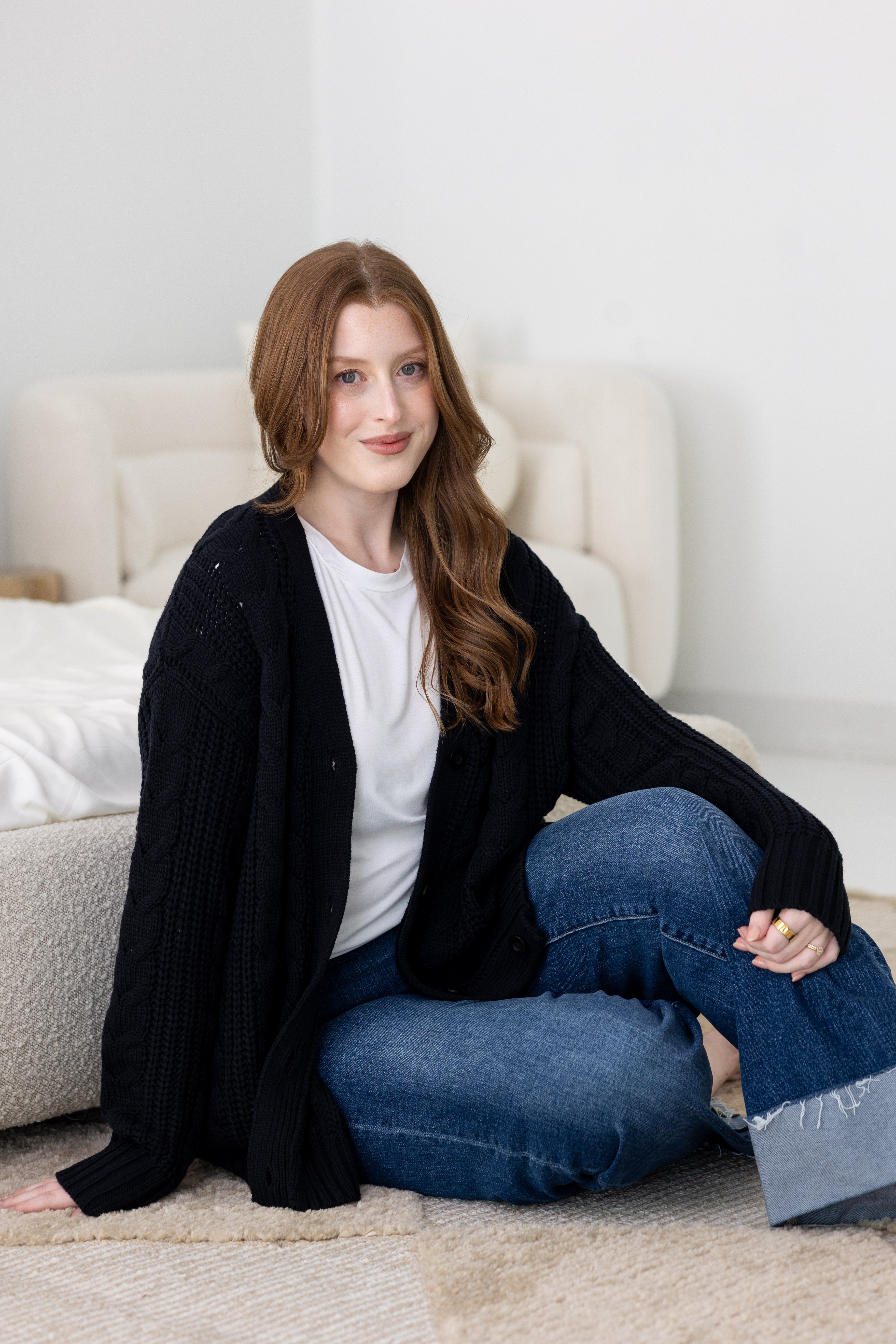 Woman sitting on a couch wearing a black cardigan and blue jeans in a home setting.