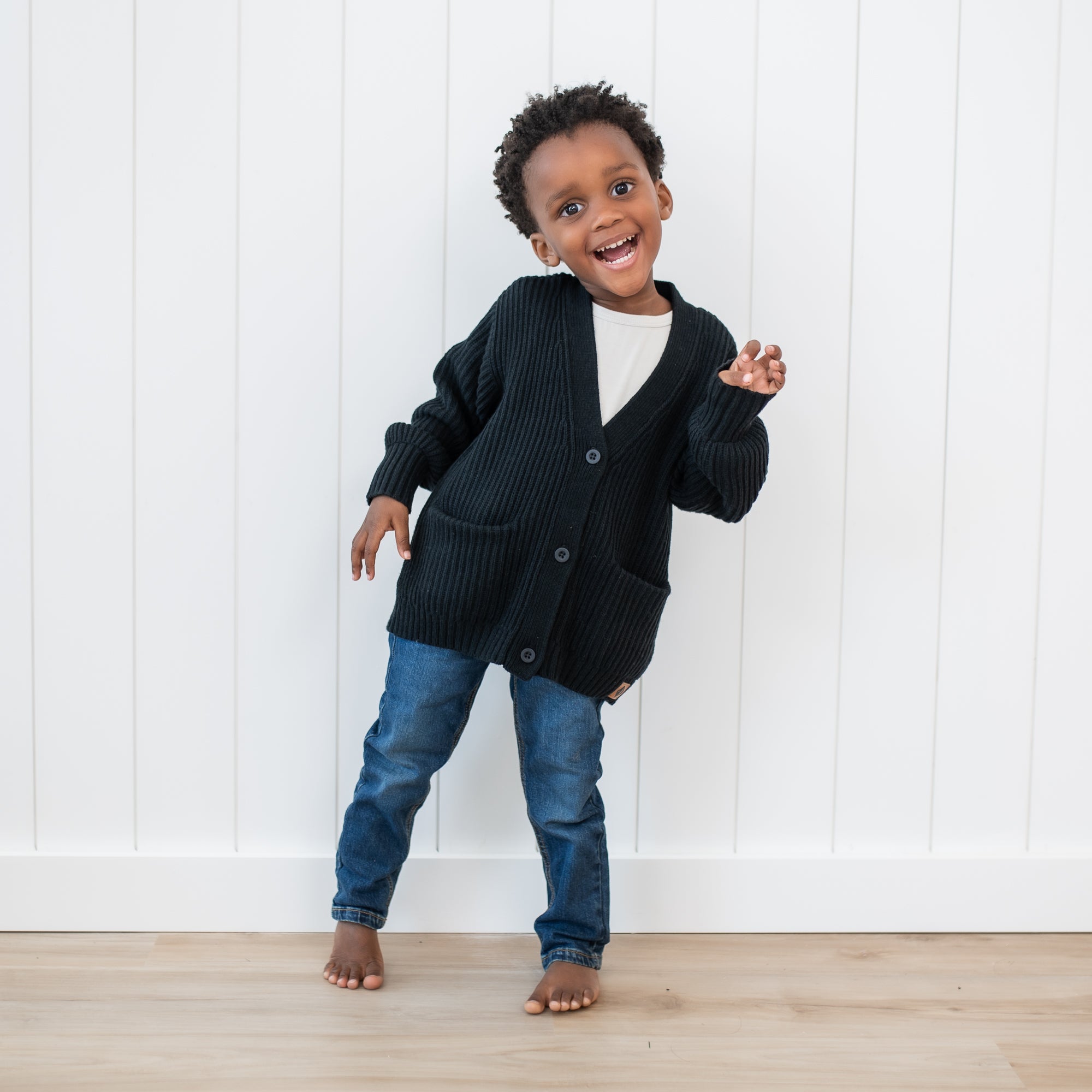 Toddler wearing the Chunky Knit Oversized Cardigan in Midnight with medium wash jeans in front of a white paneled wall