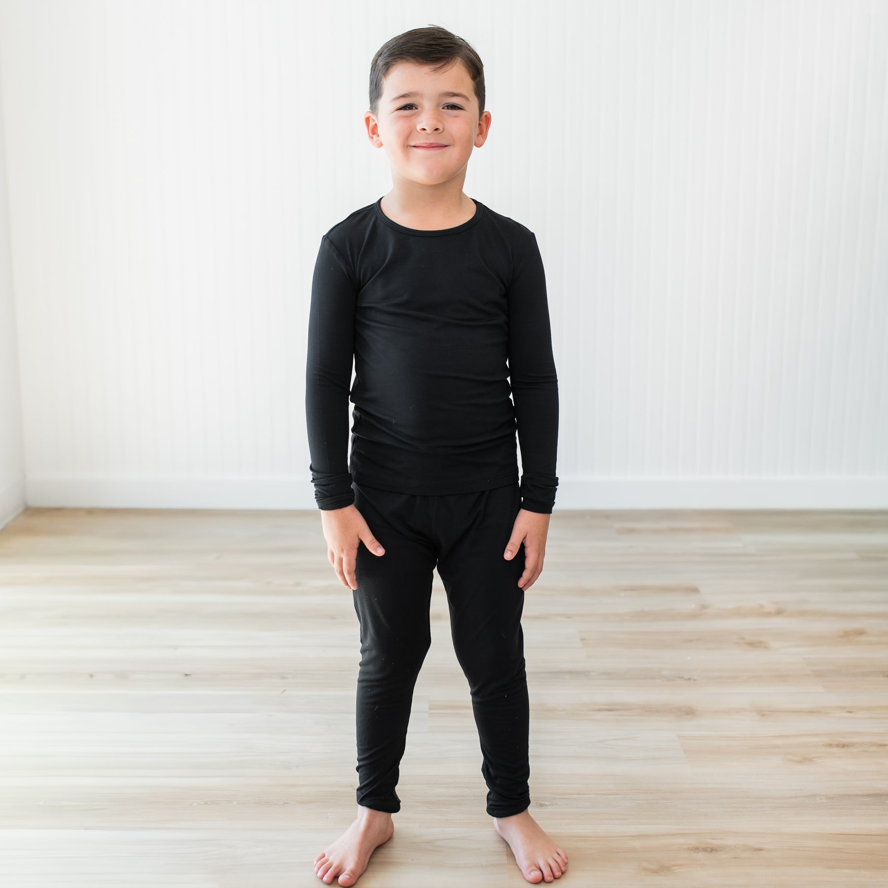 Child wearing a black long-sleeve shirt and pants pajama set standing on a wooden floor with a white wall background