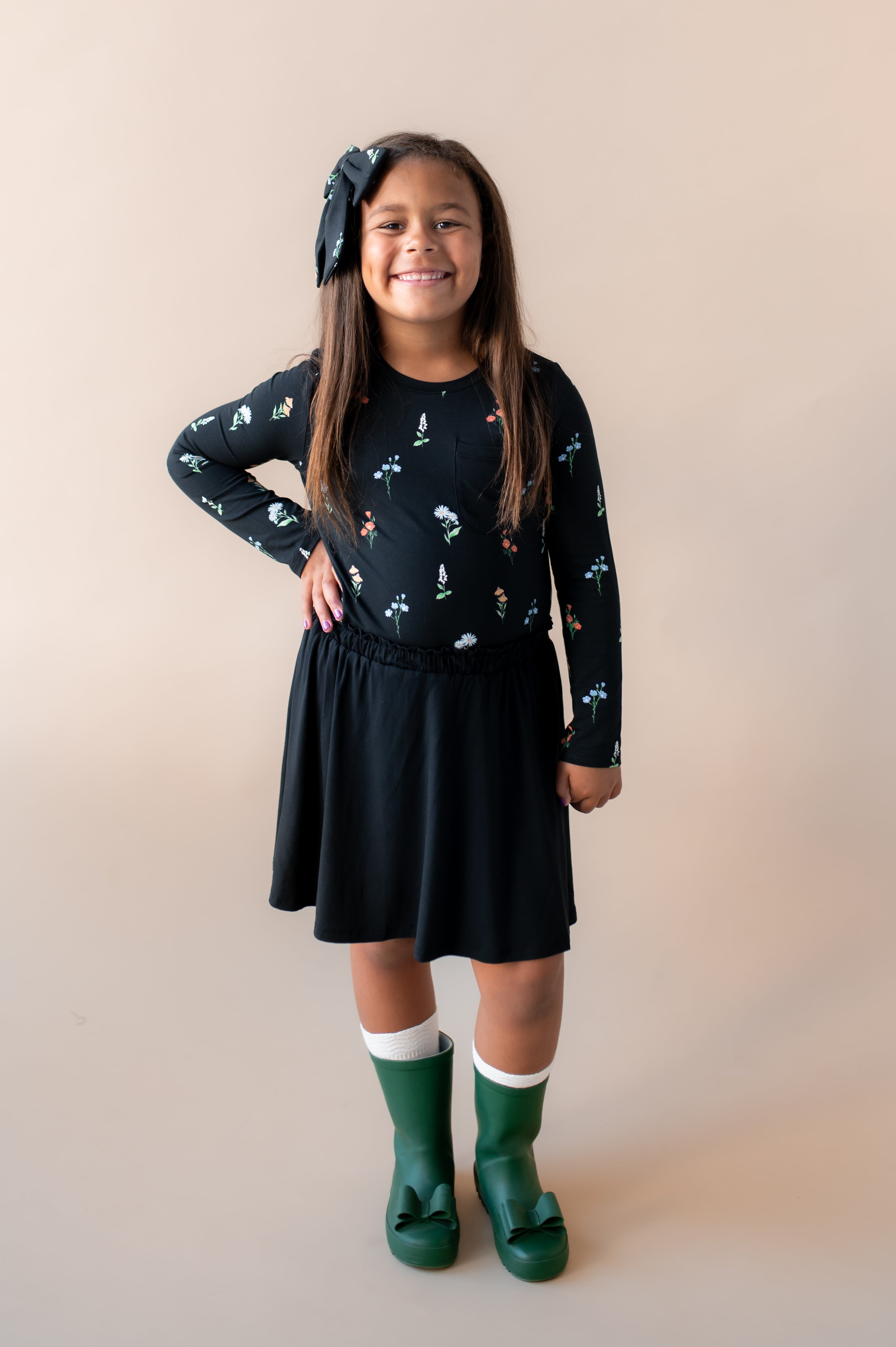 Girl posing in Long Sleeve Toddler Crew Neck Tee in Midnight Wildflower print over black skort and green shoes along with a similar print large alligator clip bow on a beige background