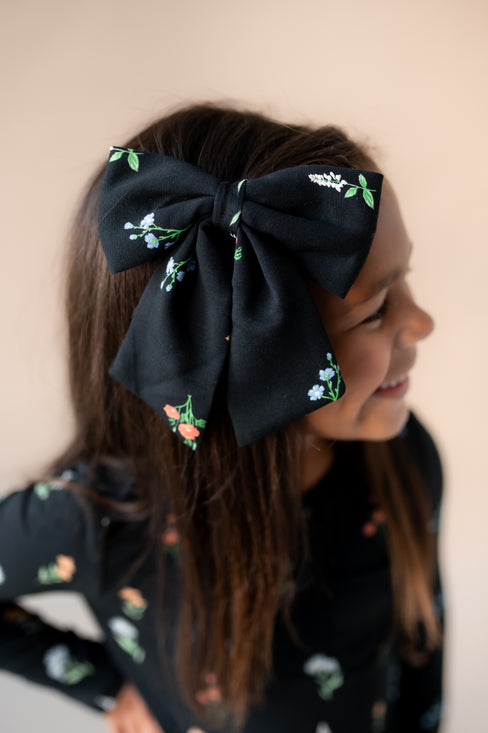 Girl wearing Large Alligator Clip Bow in Midnight Wildflower print in hair wearing a similar print dress on a beige background