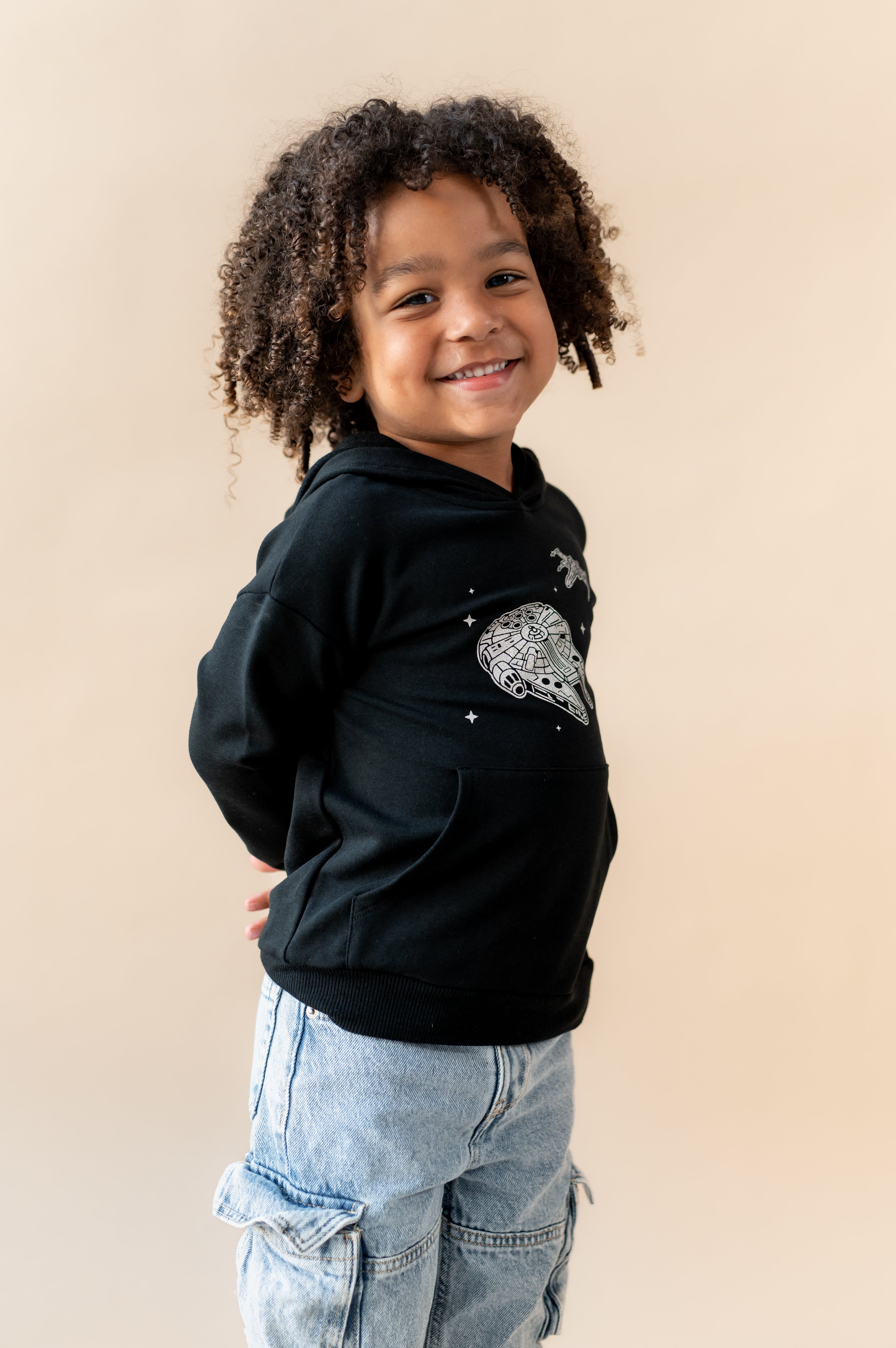 Boy smiling in Black Hoodie with Millennium Falcon™ print in center over a blue jeans on a beige background
