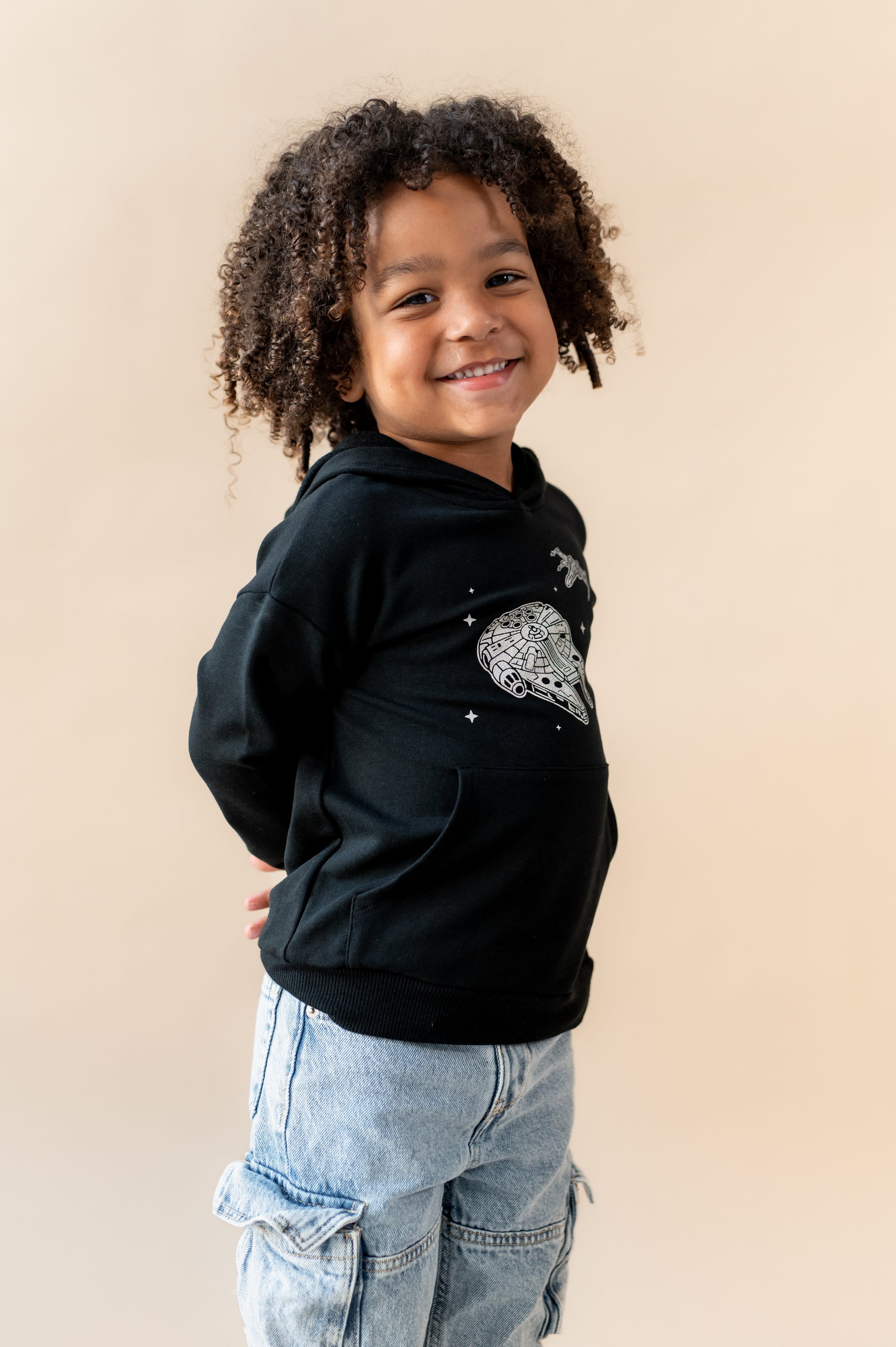 Boy smiling in Black Hoodie with Millennium Falcon™ print in center over a blue jeans on a beige background