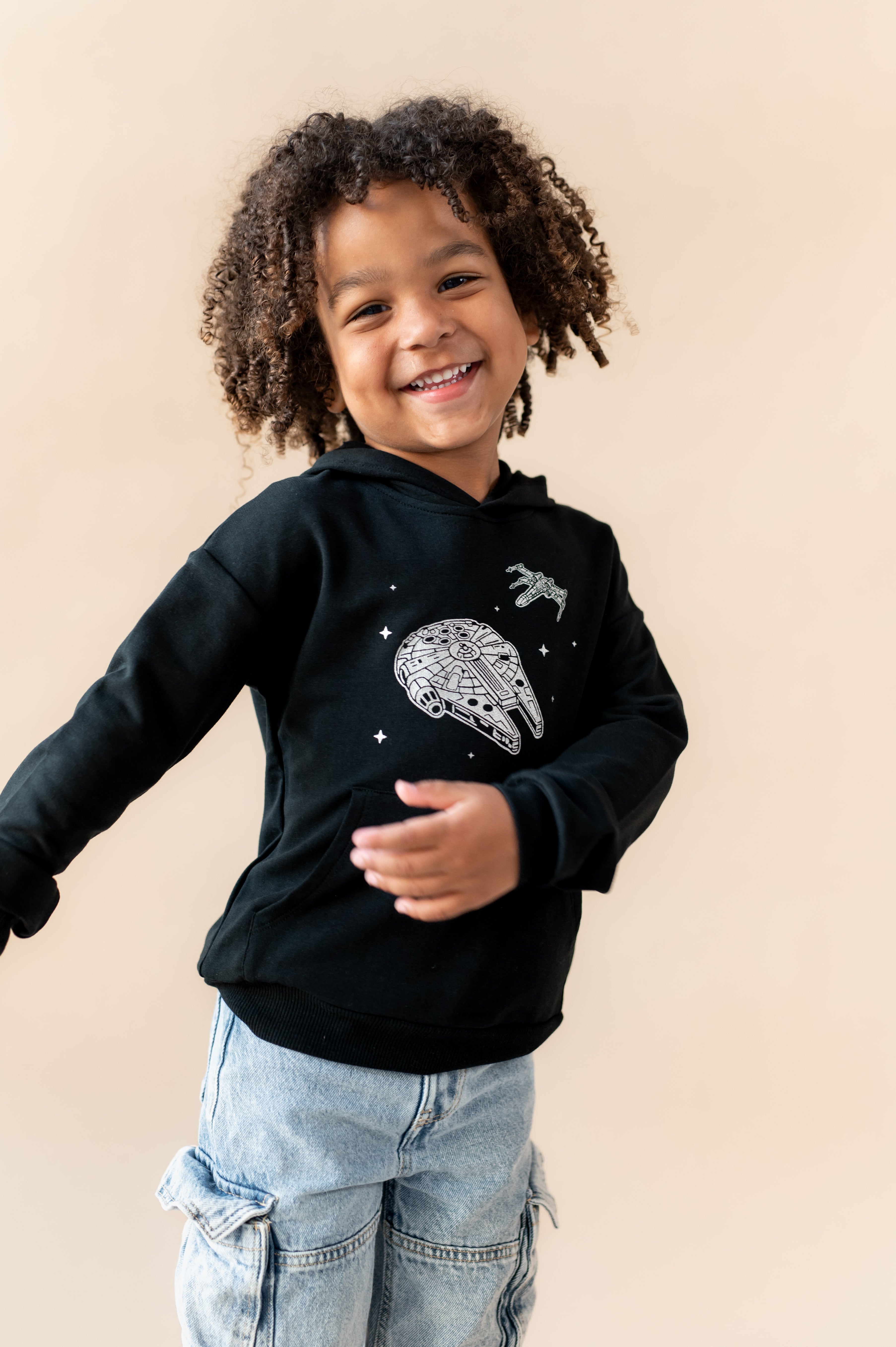 Boy posing in Black Hoodie with Millennium Falcon™ print in center over a blue jeans on a beige background