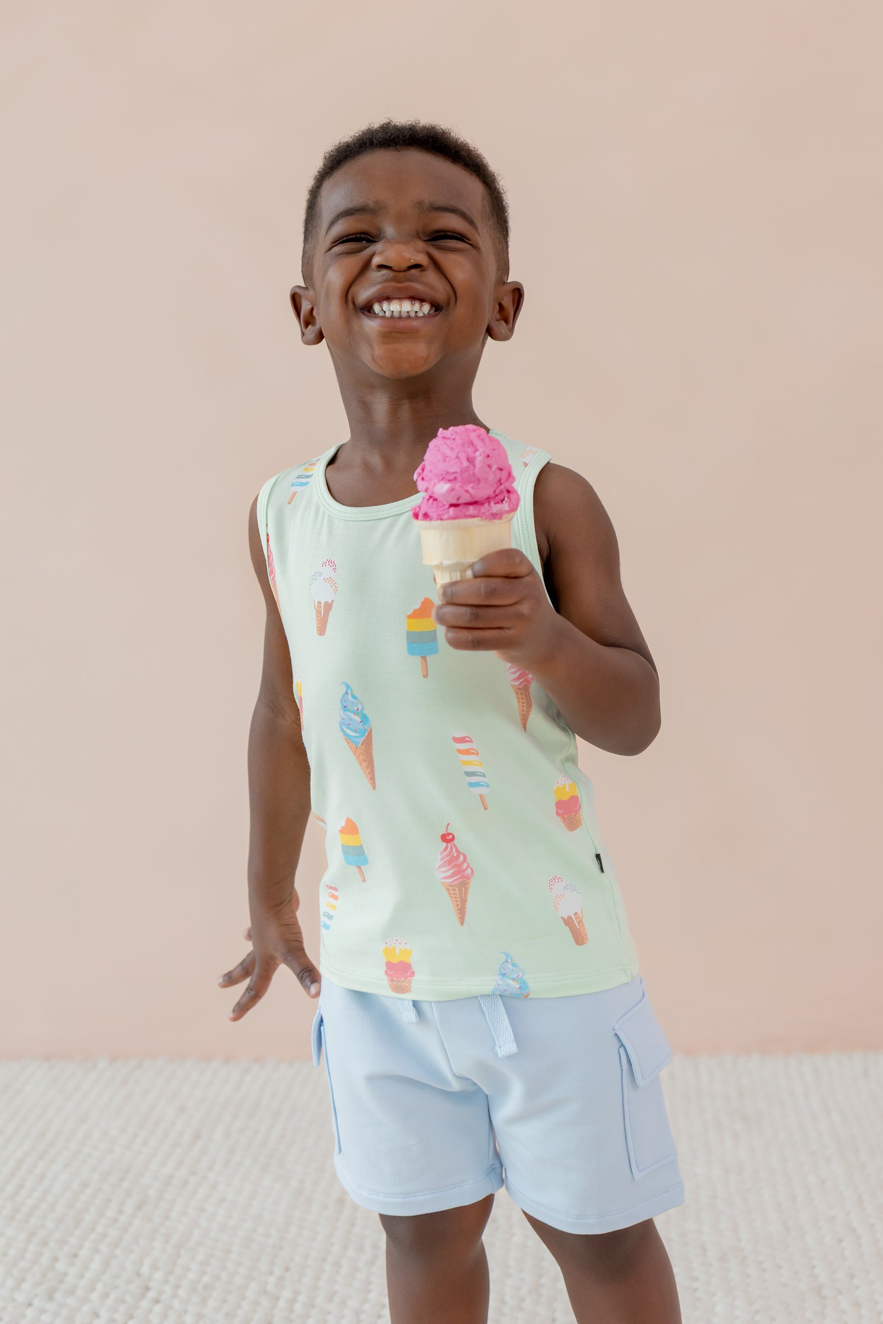 Child posing with an ice cream cone wearing a Toddler Tank Top in Mint Ice Cream print over blue shorts on a beige background