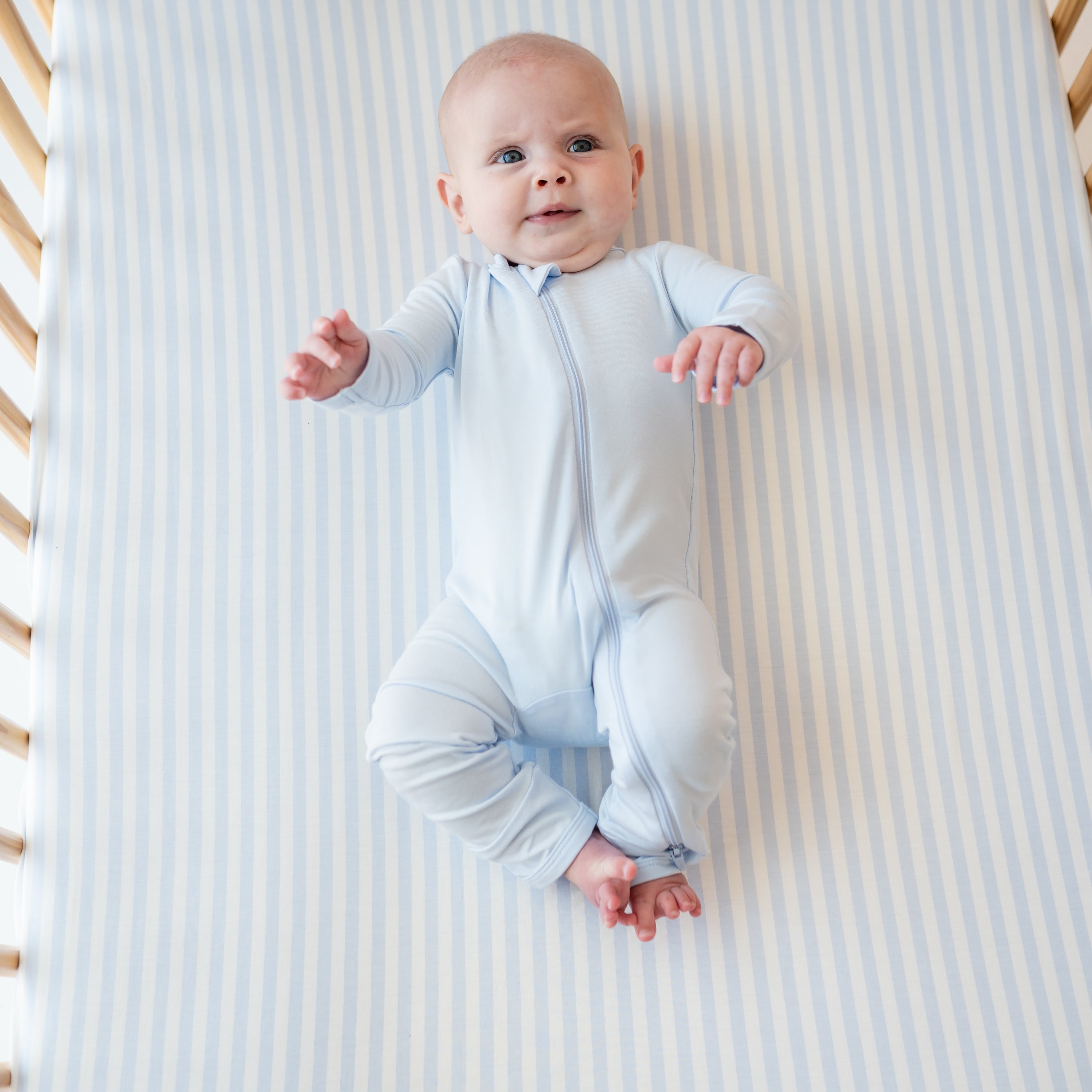 Infant wearing a Zippered Romper in Mist laying on a Crib Sheet in Small Mist Stripe 