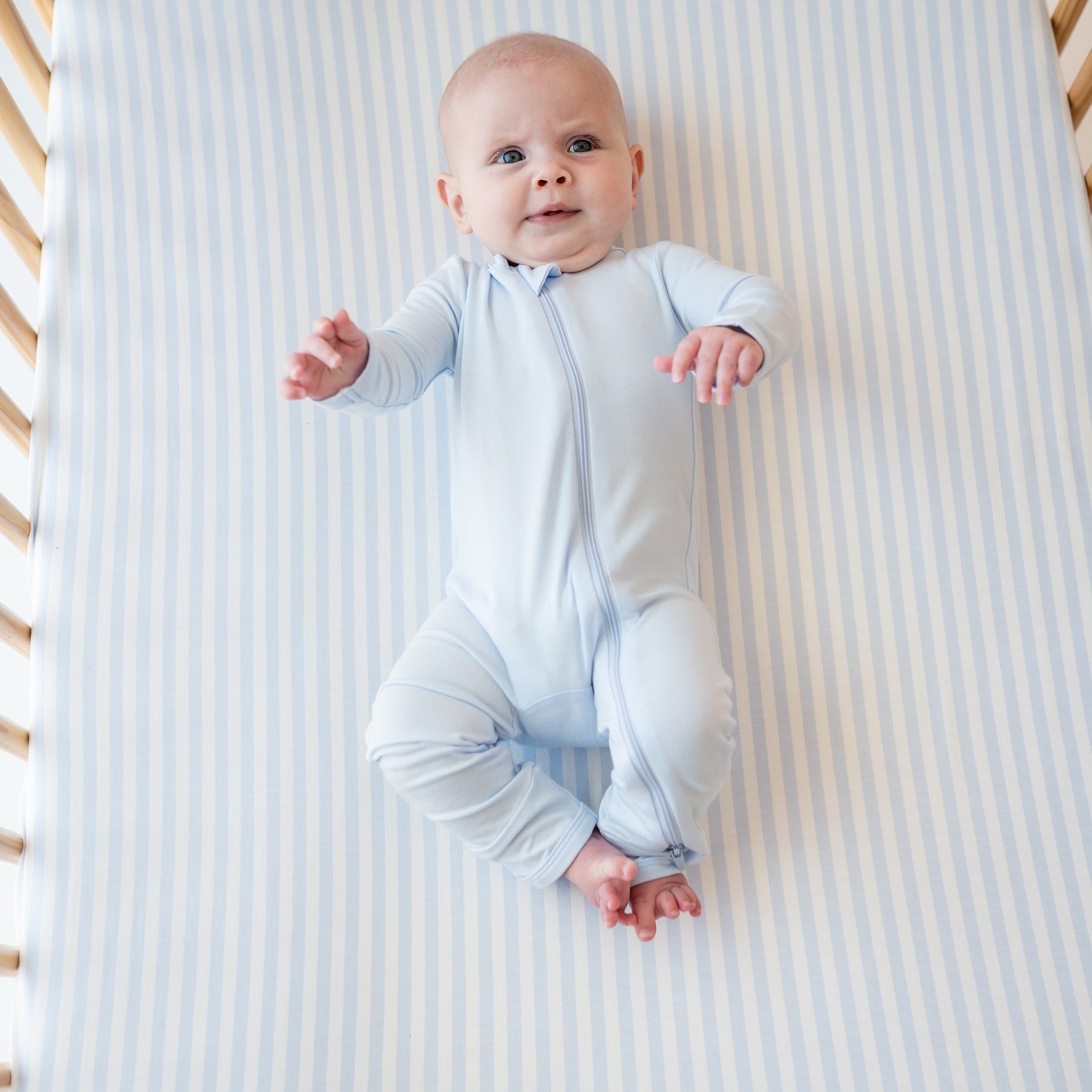 Infant wearing a Zippered Romper in Mist laying on a Crib Sheet in Small Mist Stripe