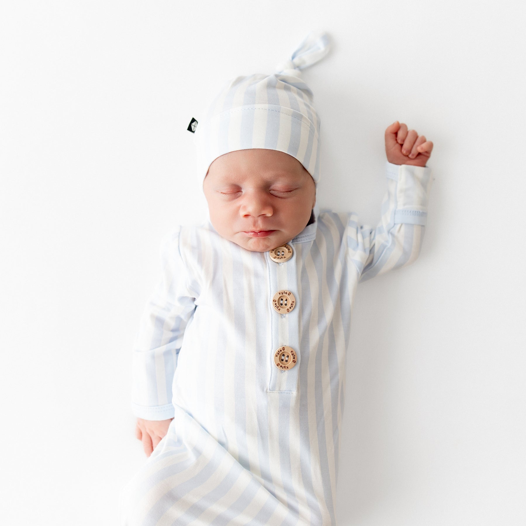 Sleeping newborn wearing the Knotted Gown with Hat Set in Small Mist Stripe laying on a white surface