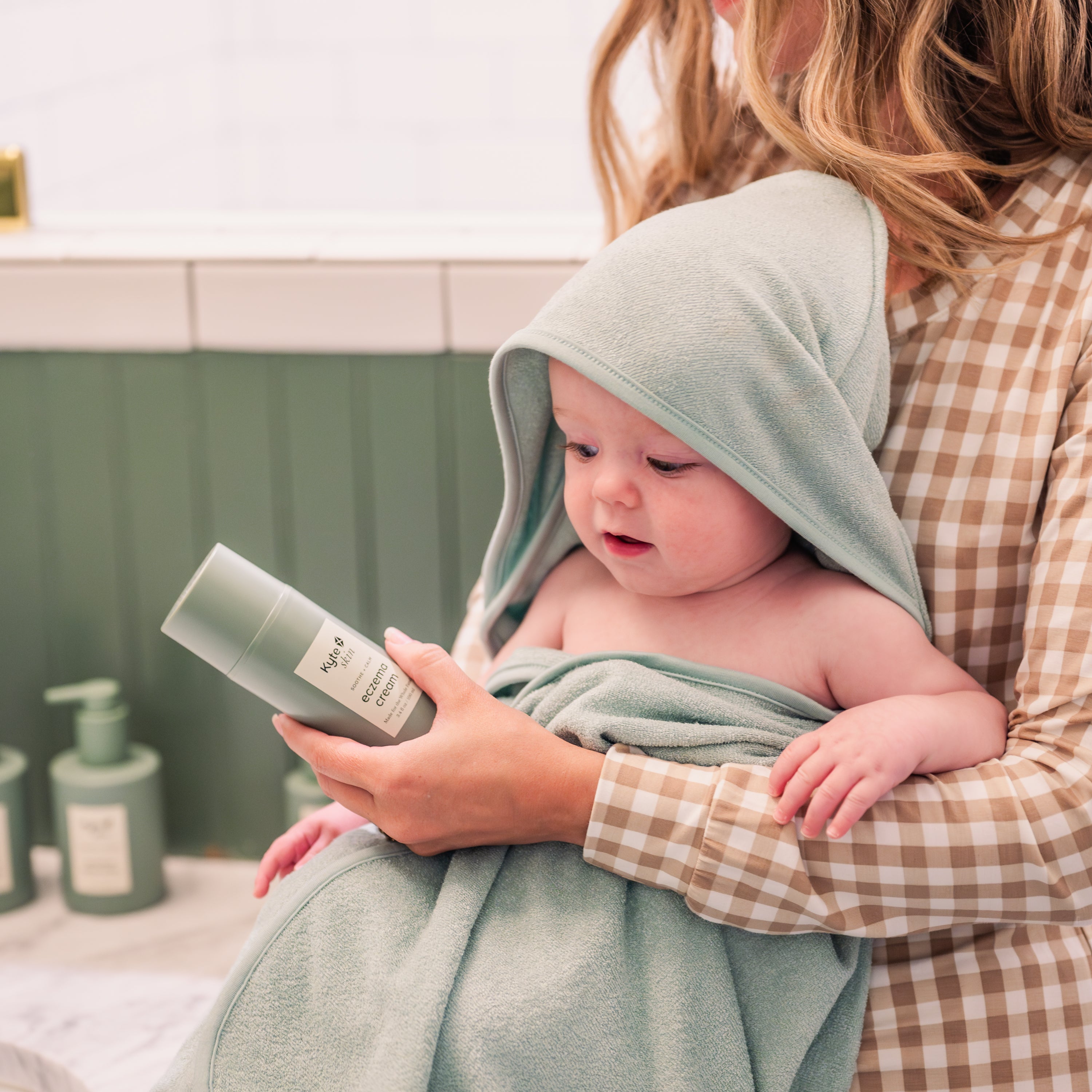 Mom holding baby wrapped in a sage green hooded bamboo terry towel holding a bottle of kyte skin eczema cream