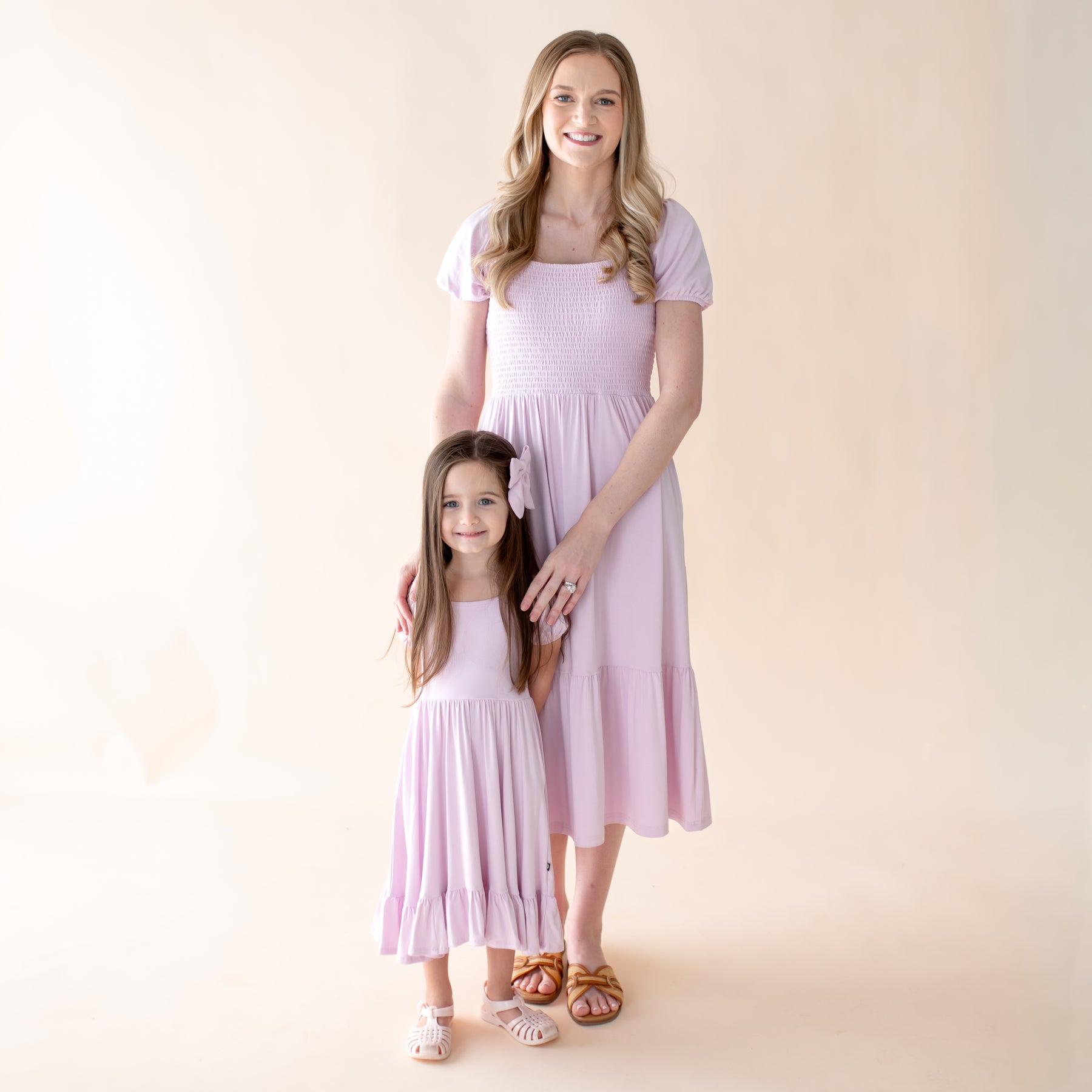 Woman and young girl wearing matching light purple dresses against a beige background