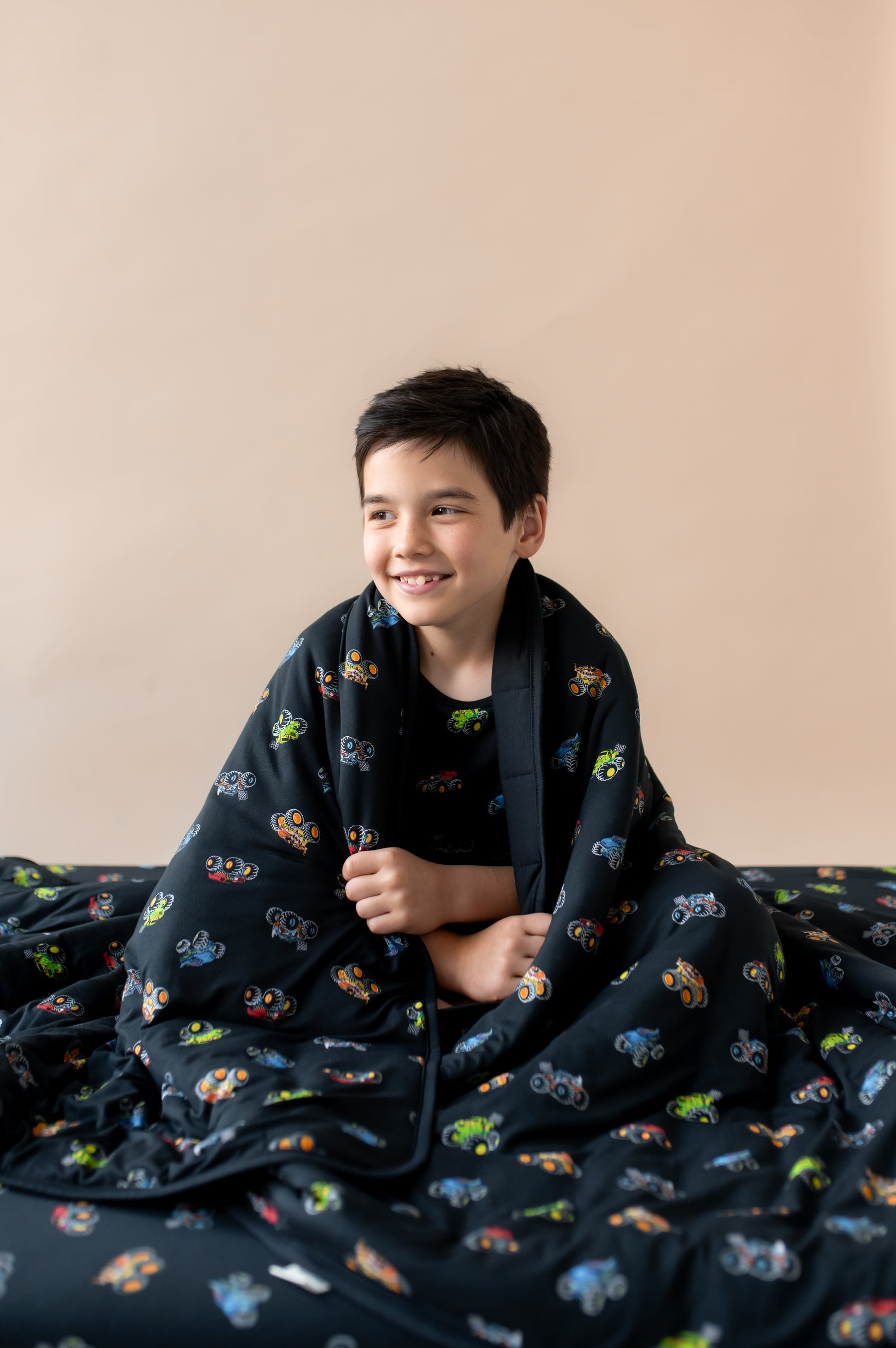 Boy wrapped in Black Youth Blanket in Monster Truck print sitting on a similar print sheet