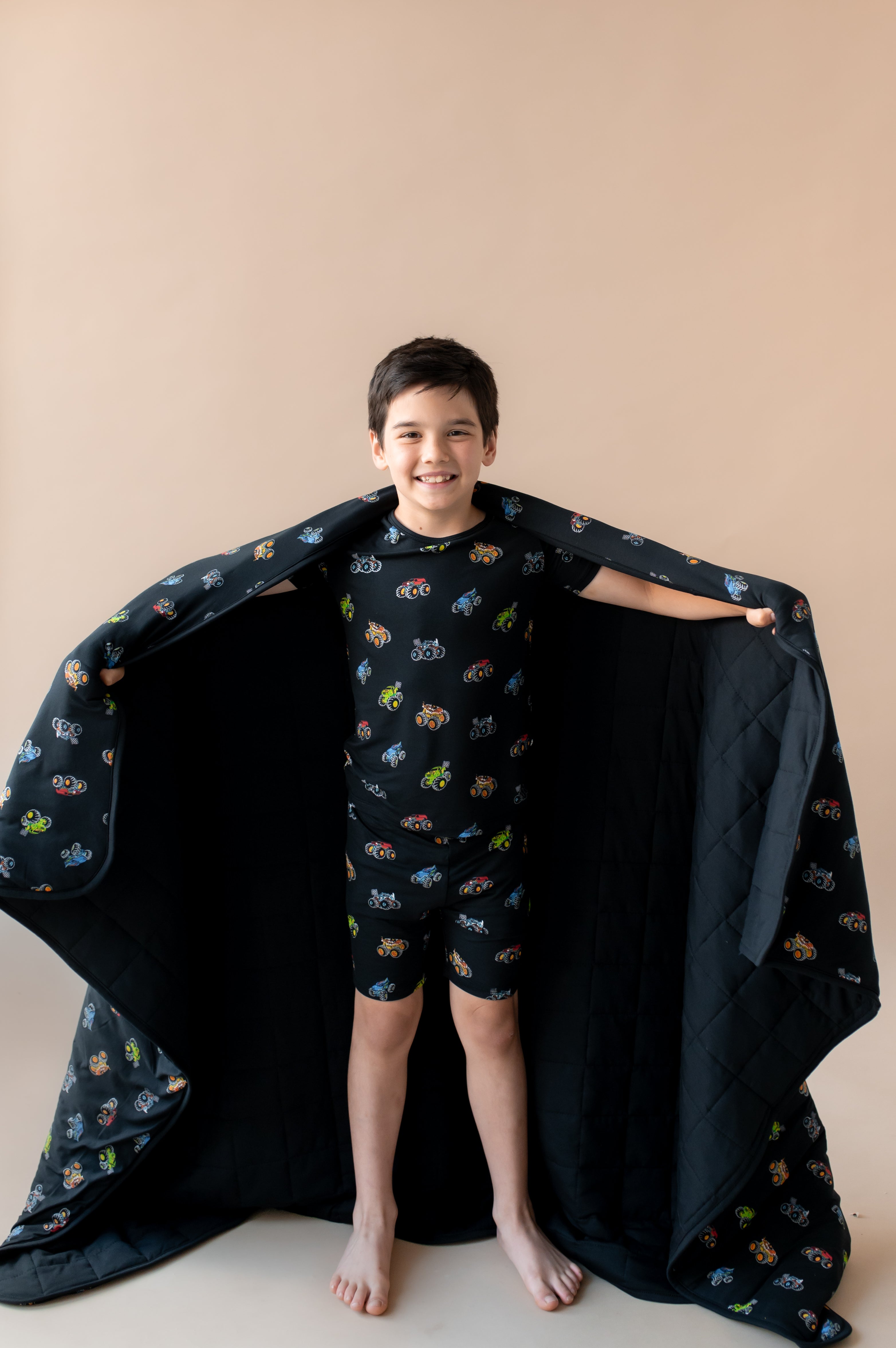 Boy holding black Youth Blanket in Monster Truck print wearing similar print short sleeve pajama set on a beige background