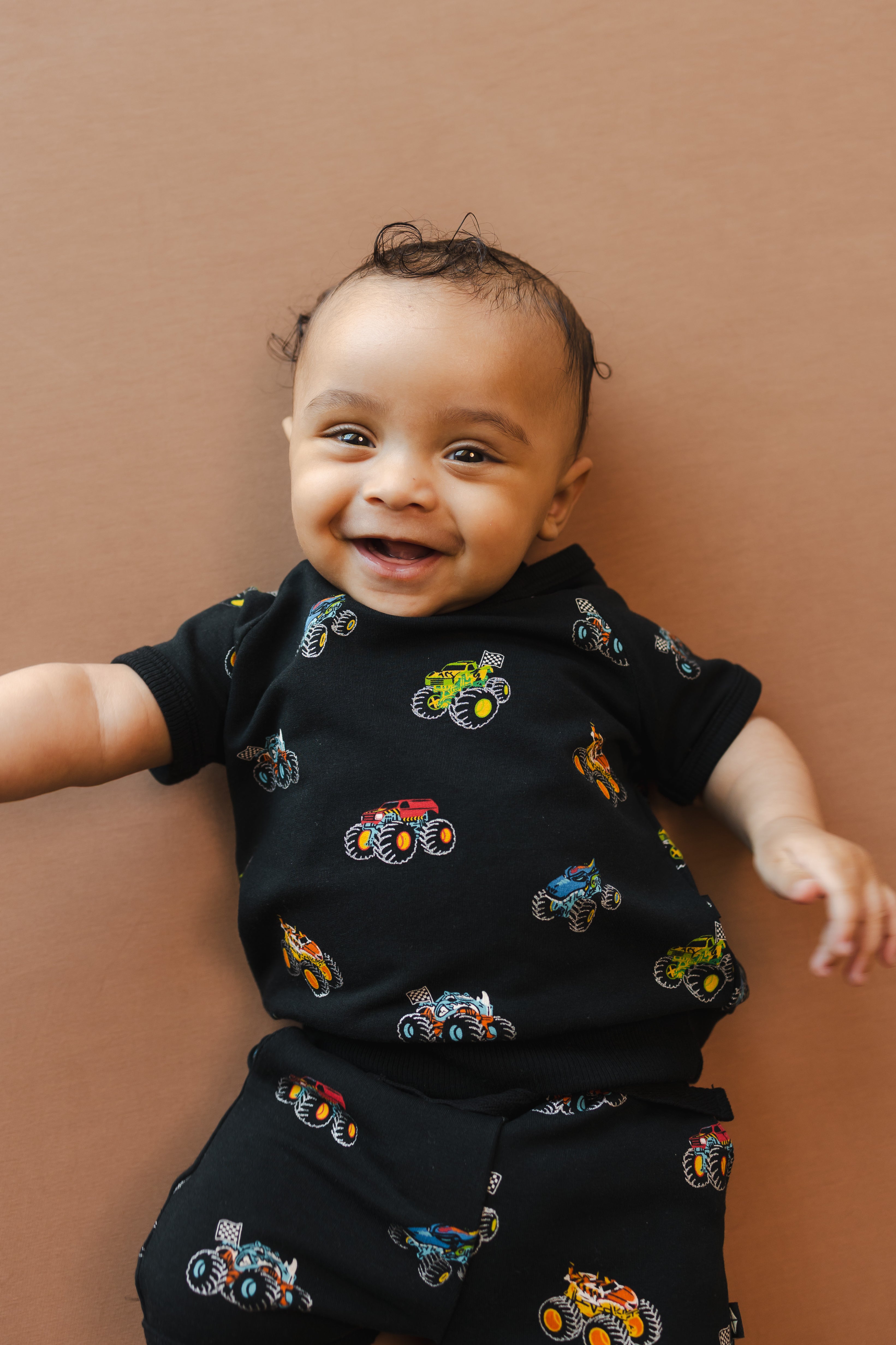Baby smiling in Black Short Sleeve Jogger Set in Monster Truck print on beige background
