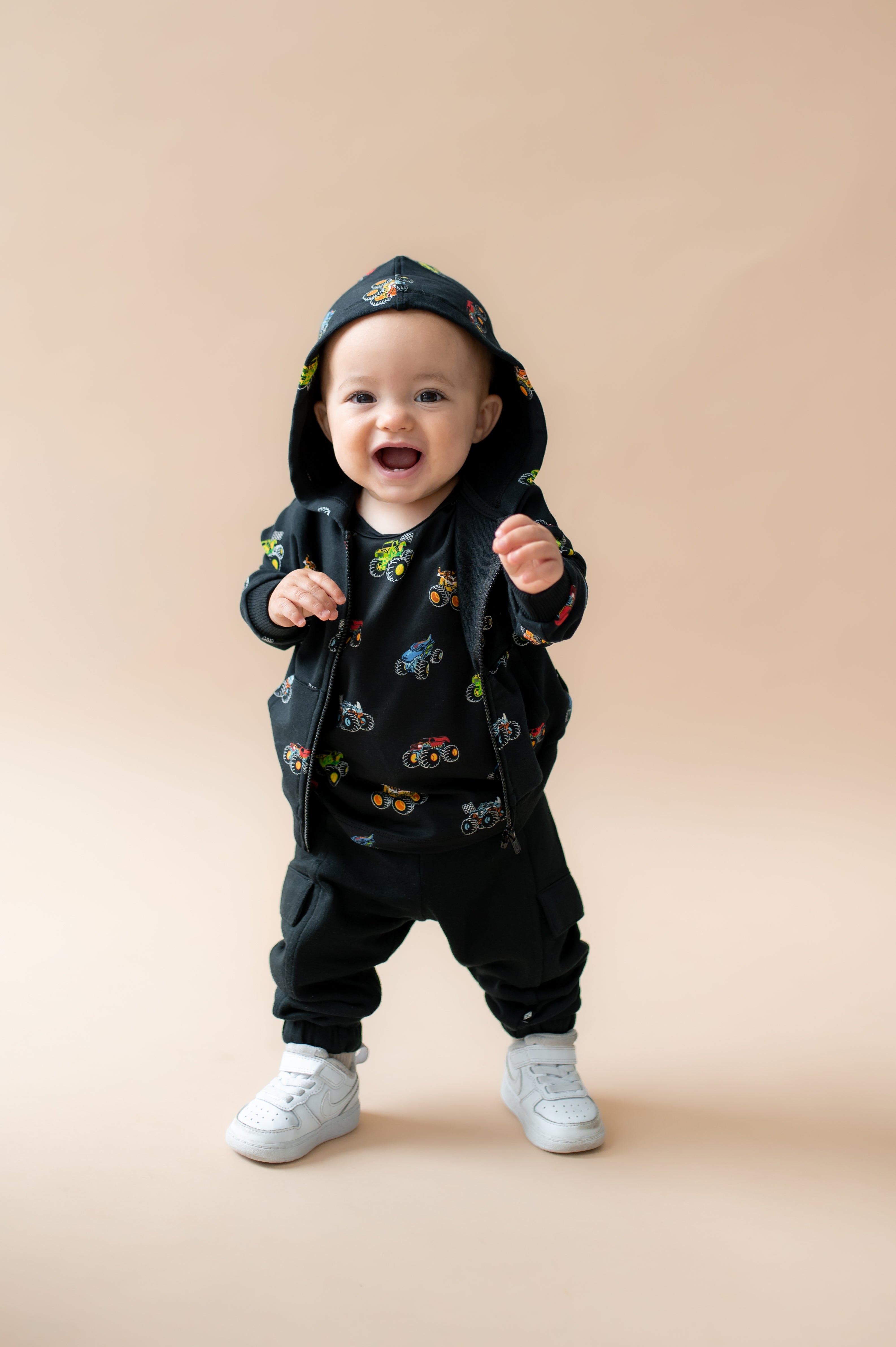 Toddler standing wearing Black Zip-Up Hoodie in Monster Truck print over black pant and white shoes on a beige background