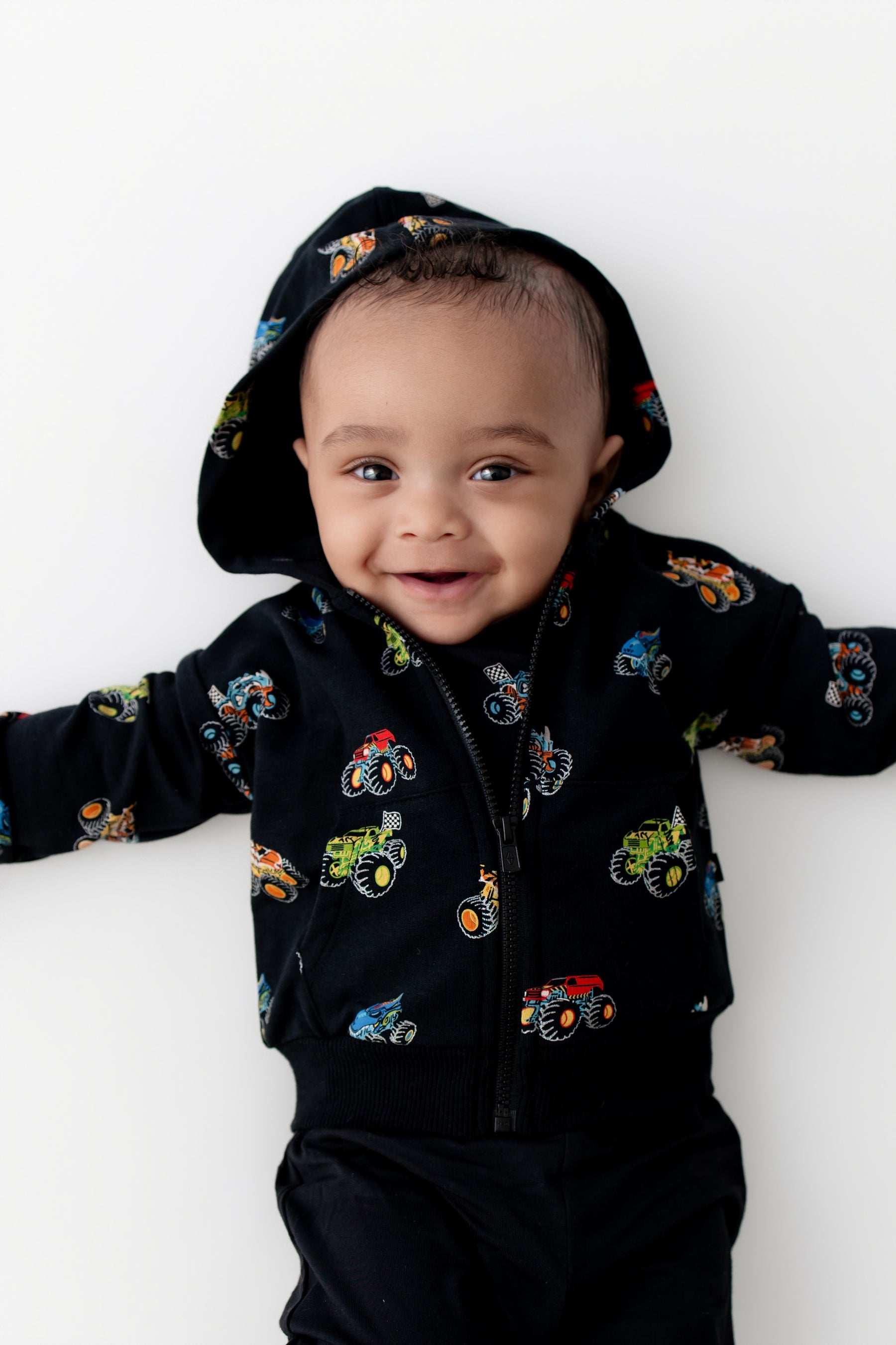 Baby in Black Zip-Up Hoodie in Monster Truck print on black pants on a white background