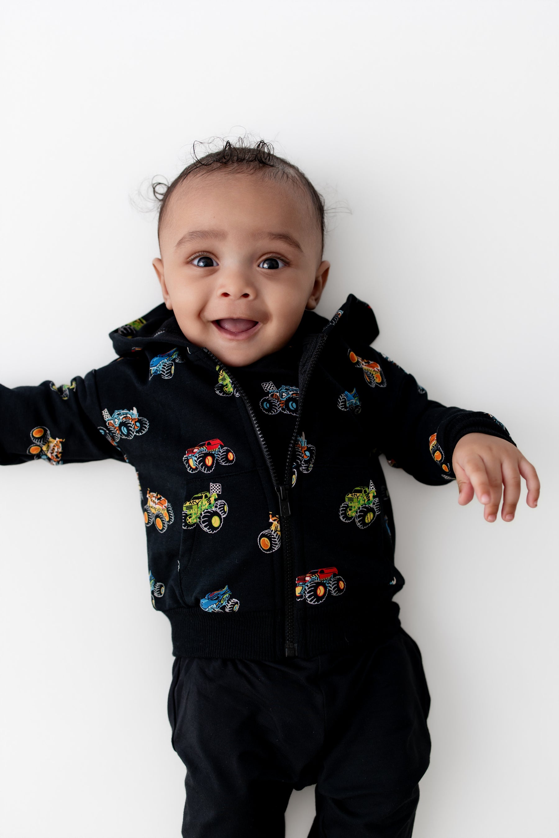 Baby smiling in Black Zip-Up Hoodie in Monster Truck print and black pant on a white background