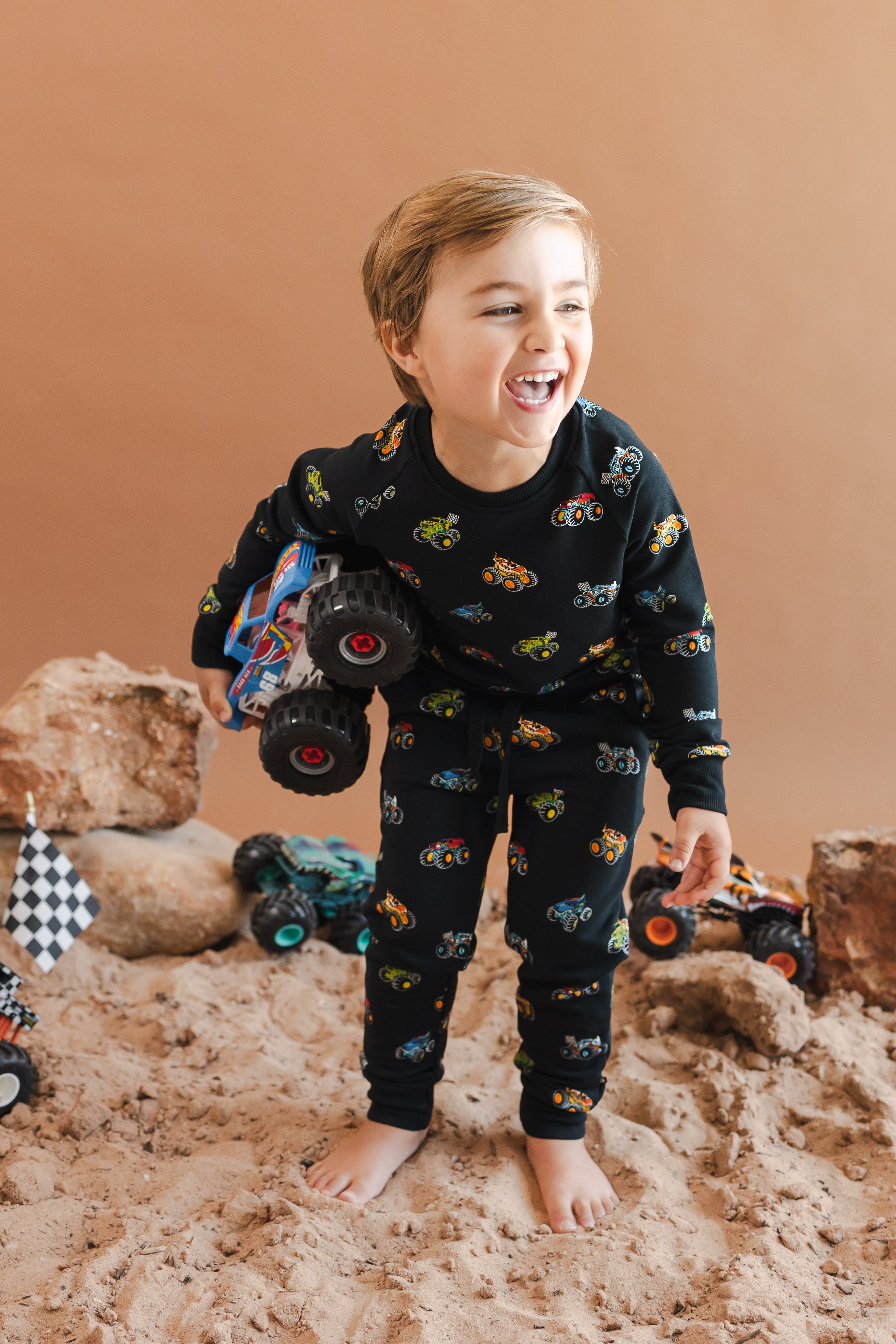 Boy holding a truck wearing black Long Sleeve Jogger Set in Monster Truck print smiling on sand and rock setup.