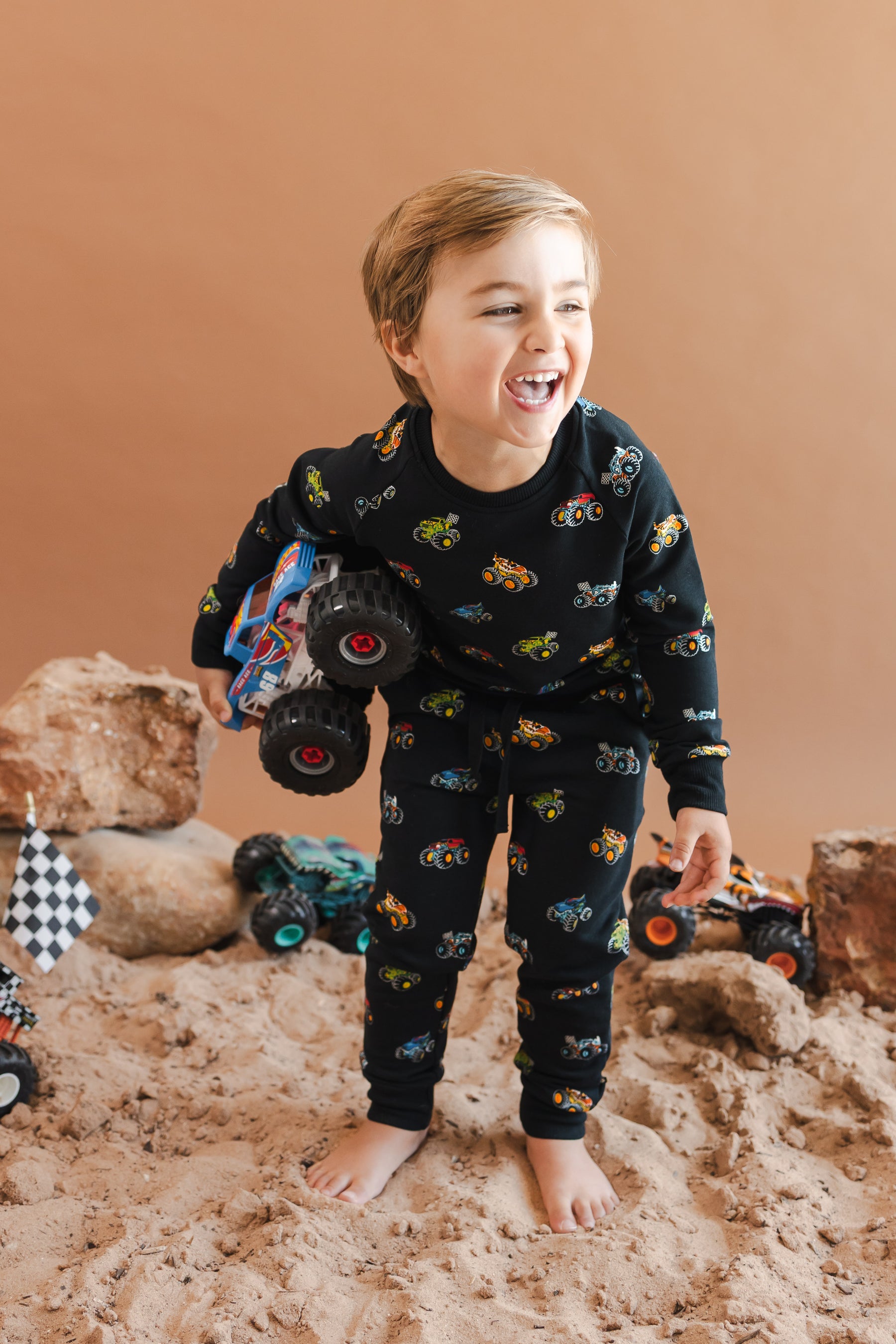 Boy holding a truck wearing black Long Sleeve Jogger Set in Monster Truck print smiling on sand and rock setup.