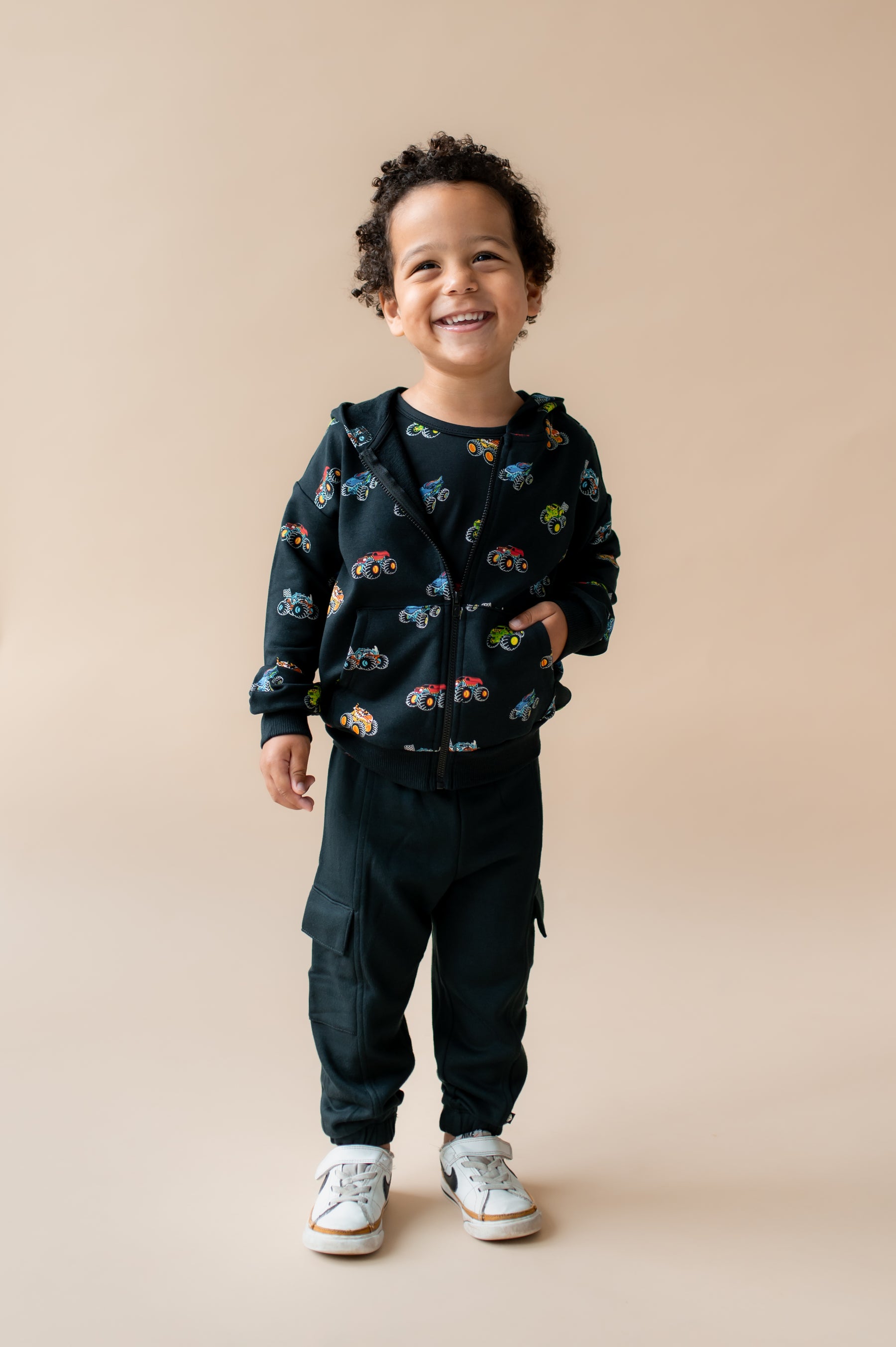 Boy smiling in Black Zip-Up Hoodie in Monster Truck print and black pant and white shoes on a beige background