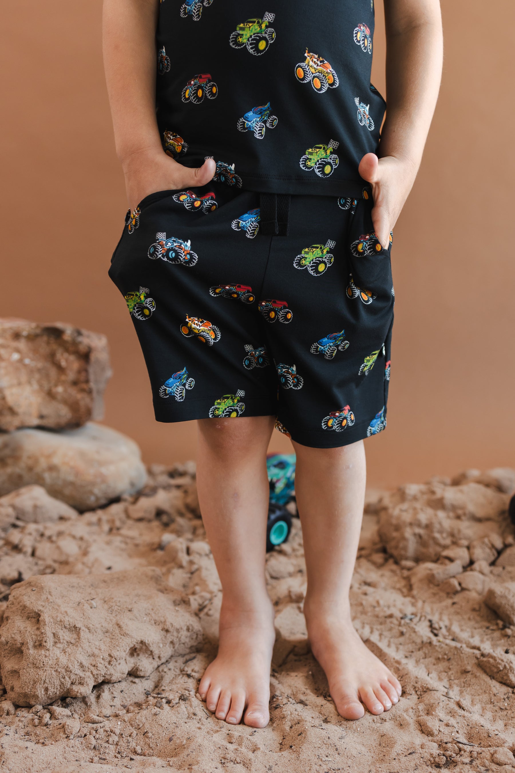 Boy wearing drawstring short with pockets in Monster Truck print standing on sand and rock with toy trucks