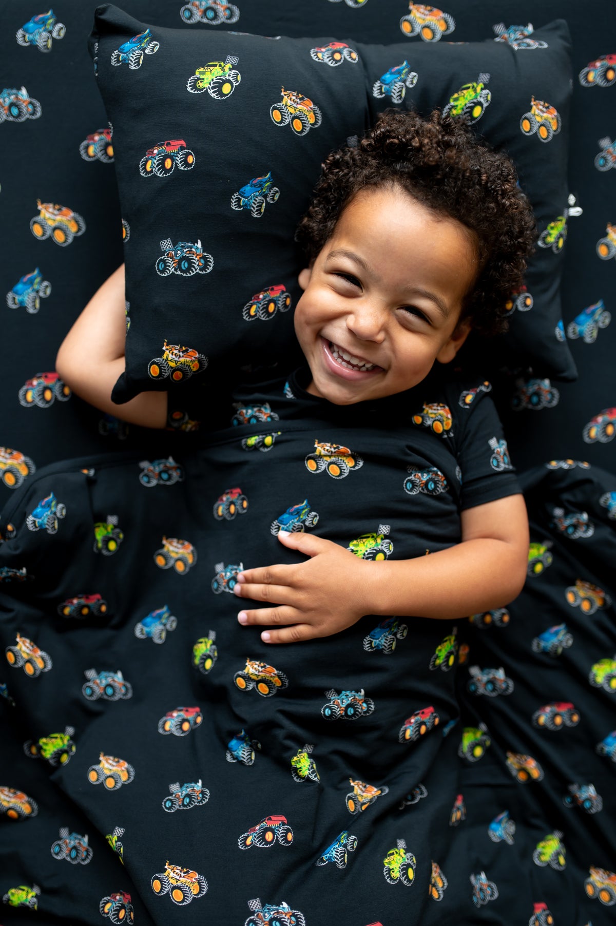Boy lying on black Toddler pillowcase and sheet with Monster Truck print under a similar print Toddler blanket