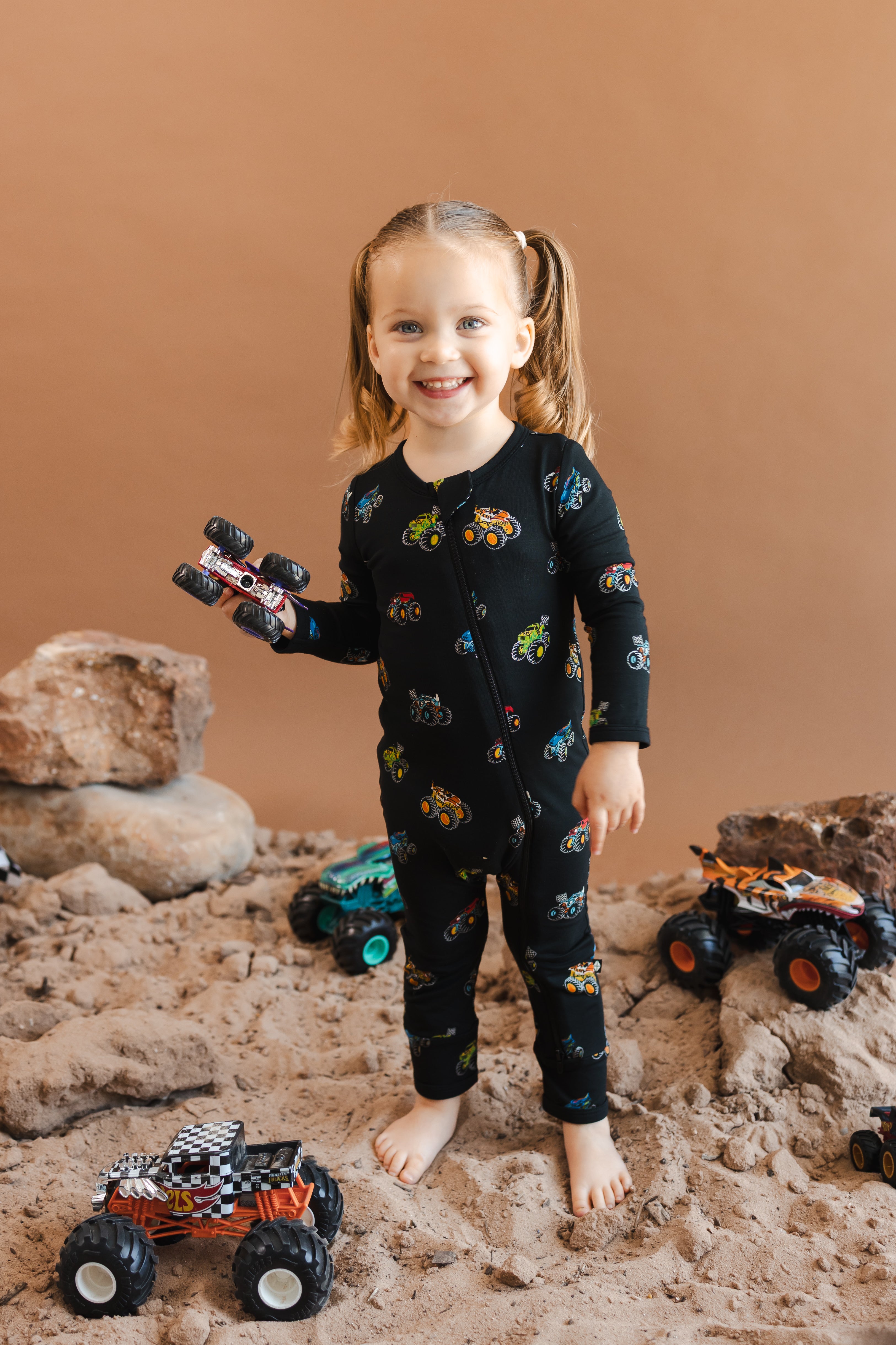 Girl holding toy truck wearing black Zippered Romper in Monster Truck print on sand and rocks with toy trucks