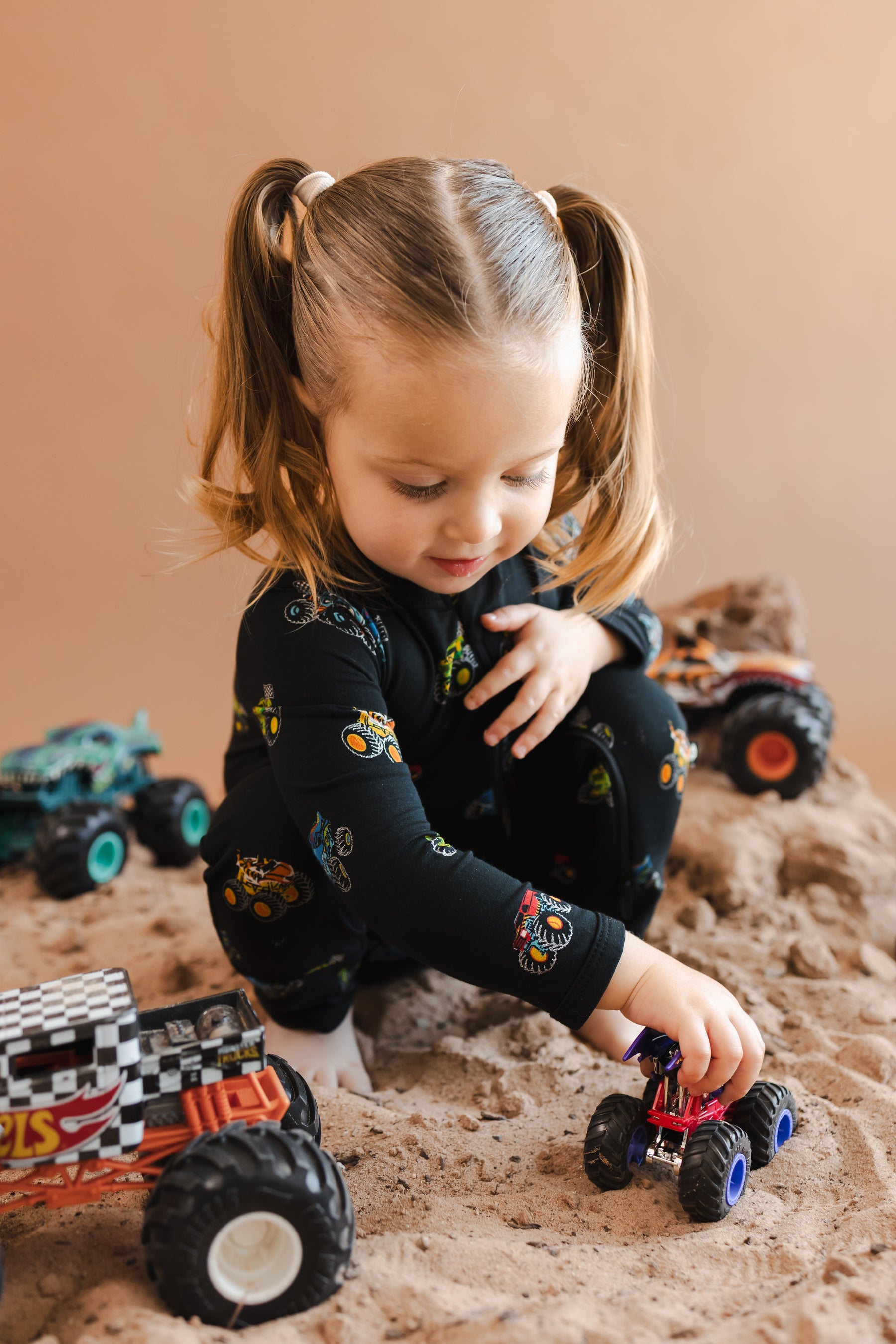 Girl playing with toy truks on sand and rocks wearing black Zippered Romper in Monster Truck print