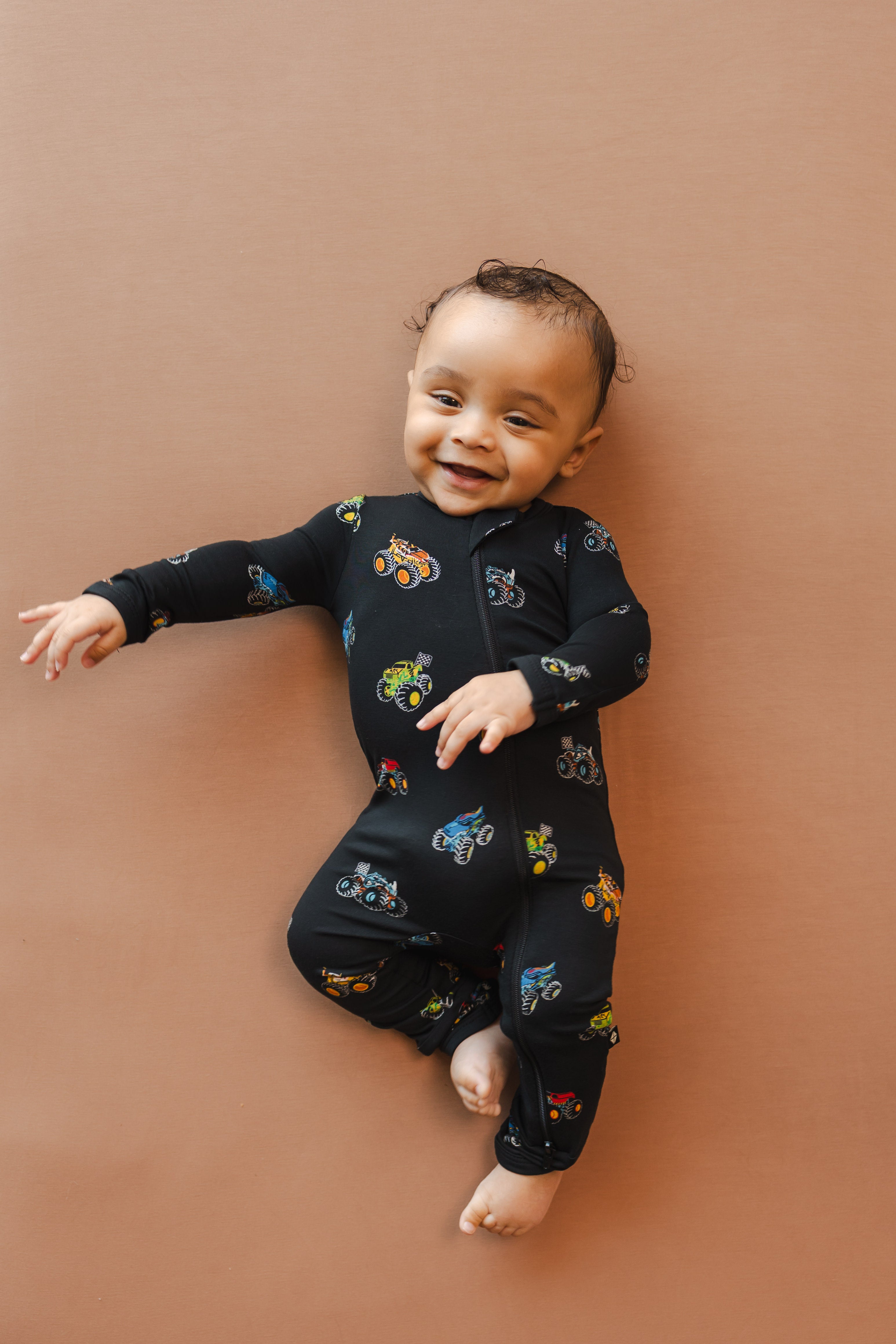 Baby wearing black Zippered Romper in Monster Truck print on beige background
