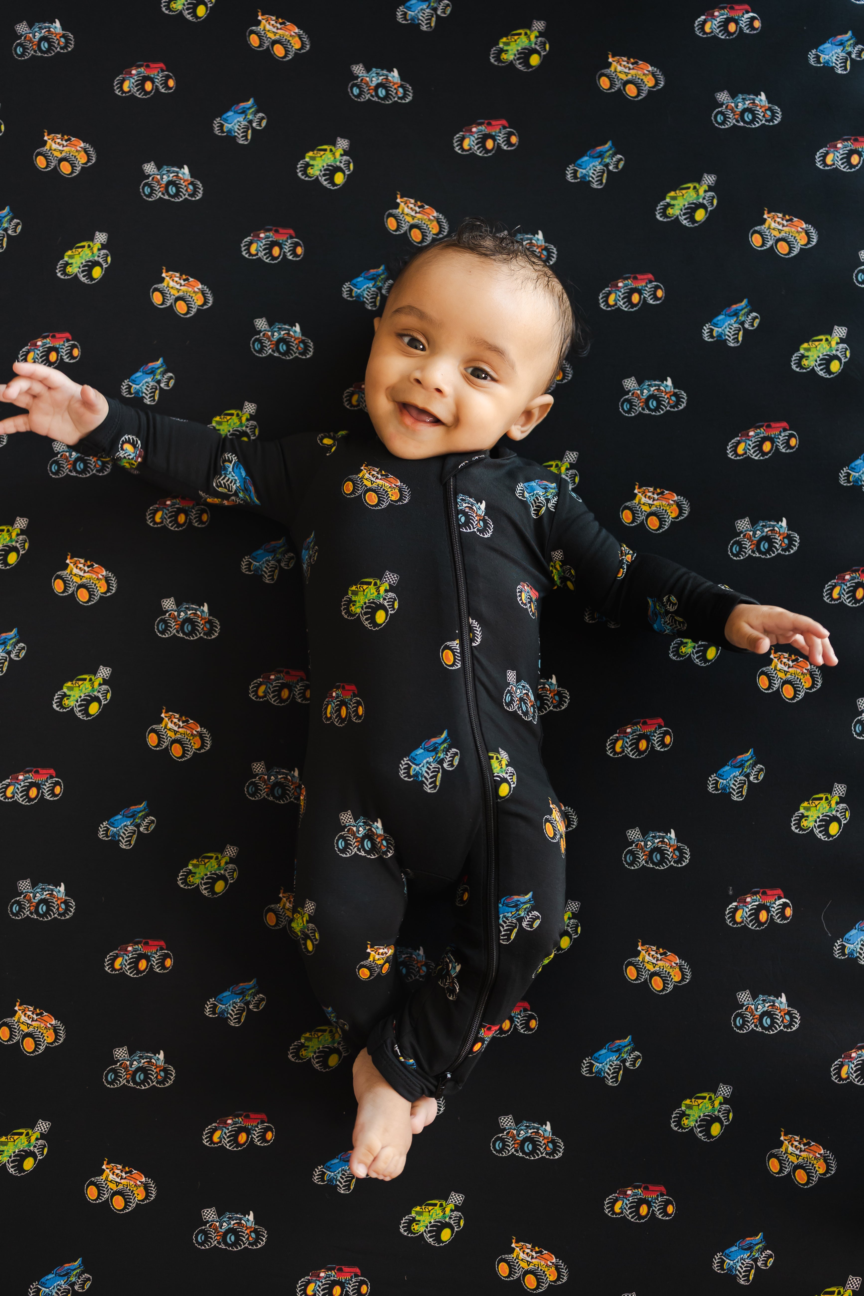 Baby smiling on black crib sheet with Monster truck print wearing similar print Zippered Romper 