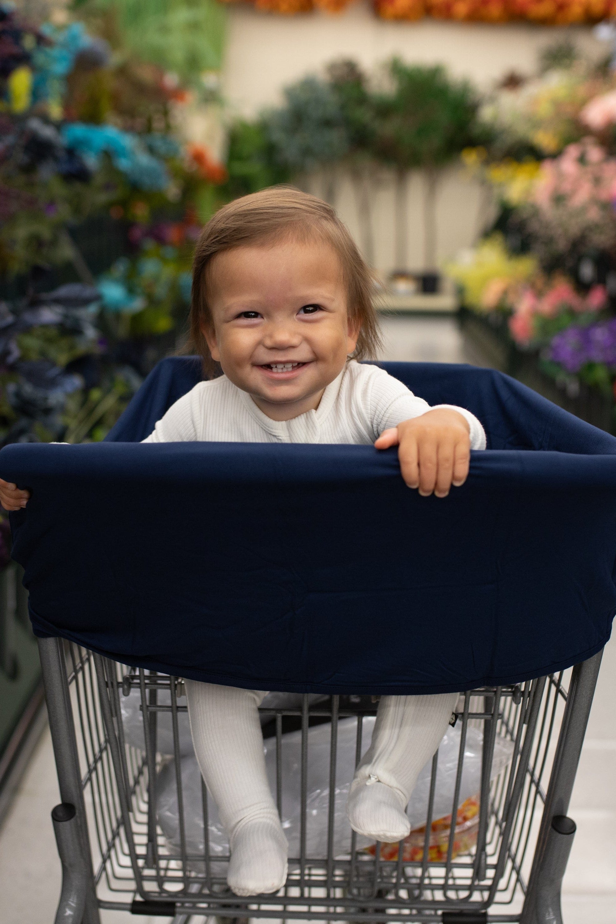 Toddler in a shopping cart with a navy blue cover in a grocery store aisle with flowers in the background