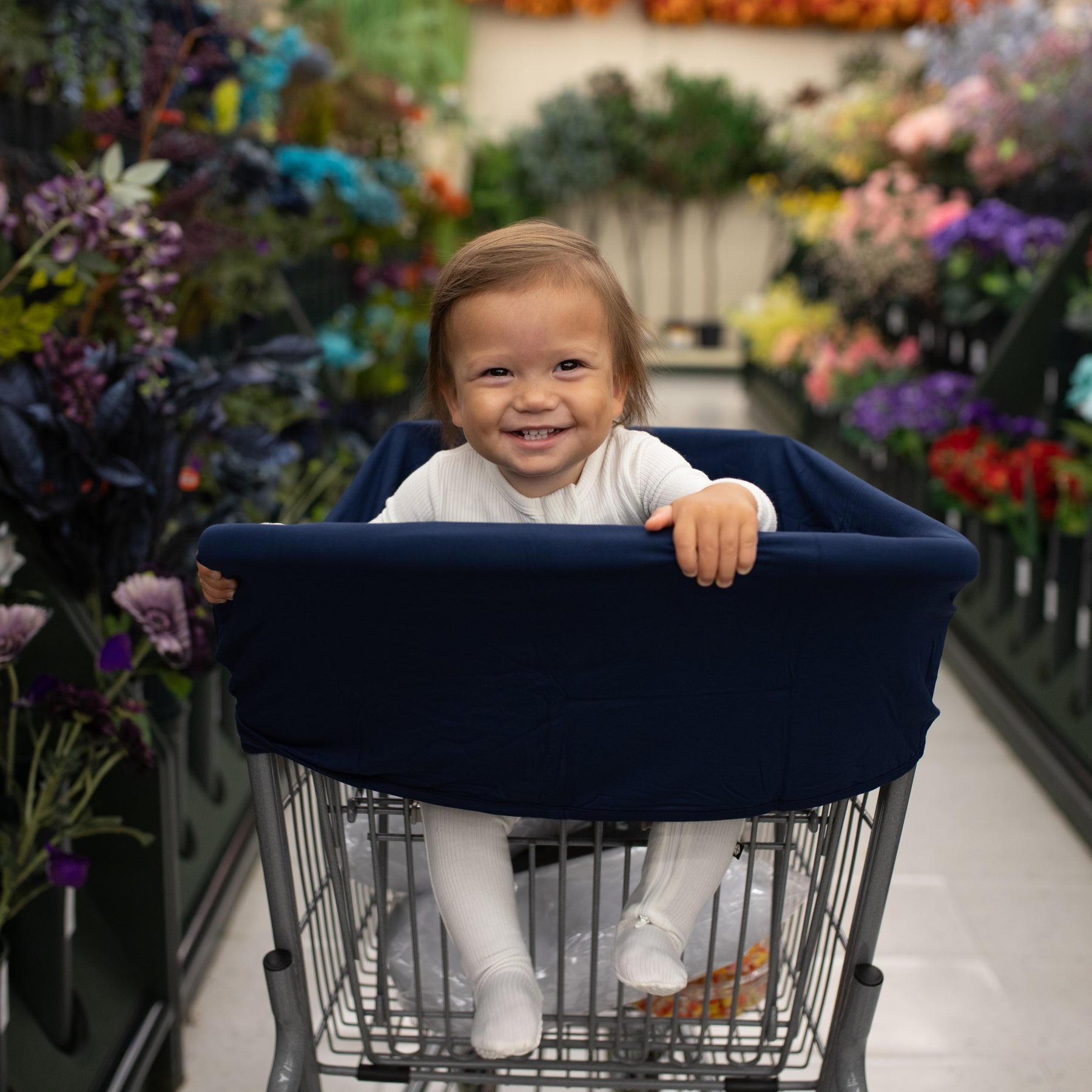 Toddler in a shopping cart with a navy blue cover in a grocery store aisle with flowers in the background