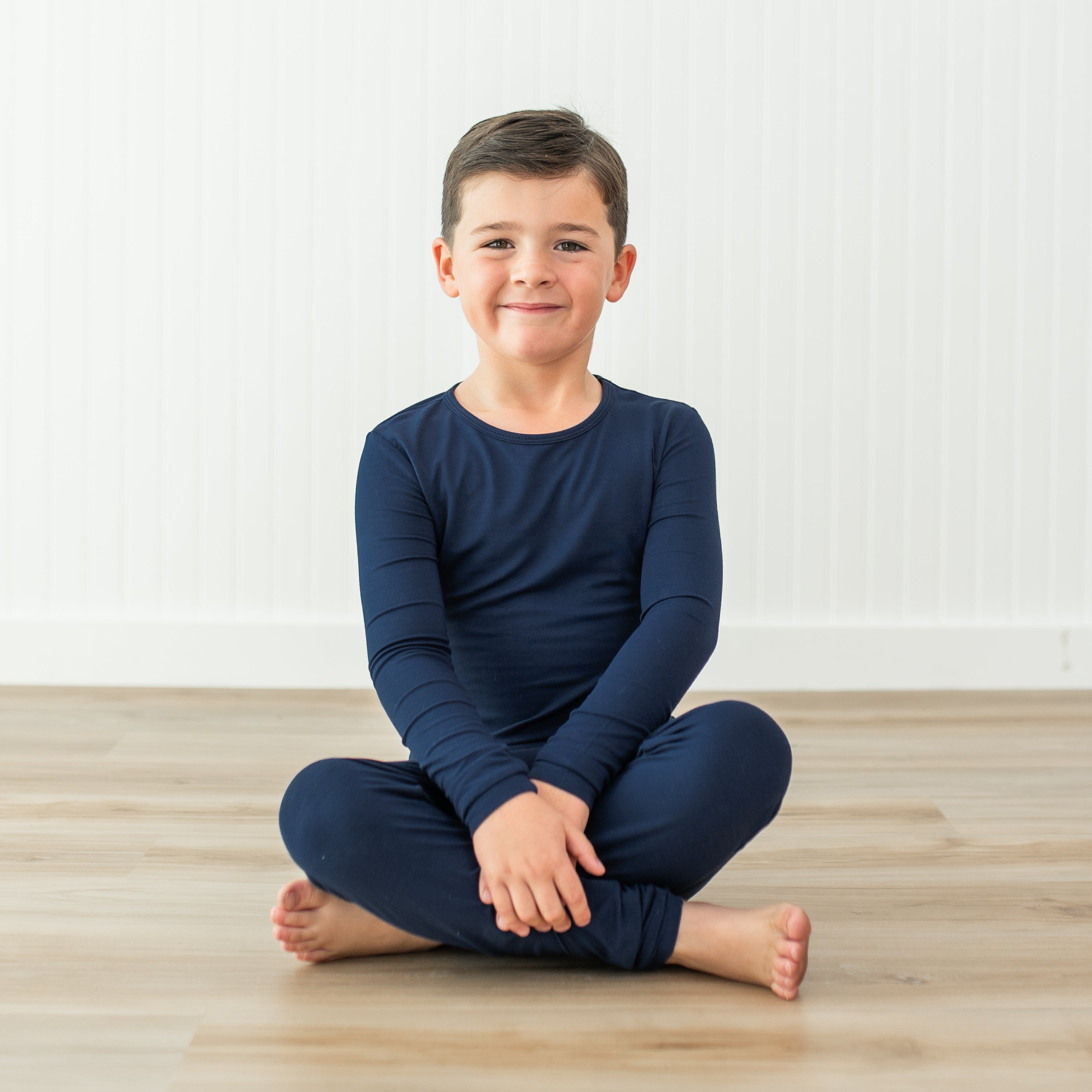 Young boy sitting on a wooden floor wearing navy blue pajamas against a white wall.