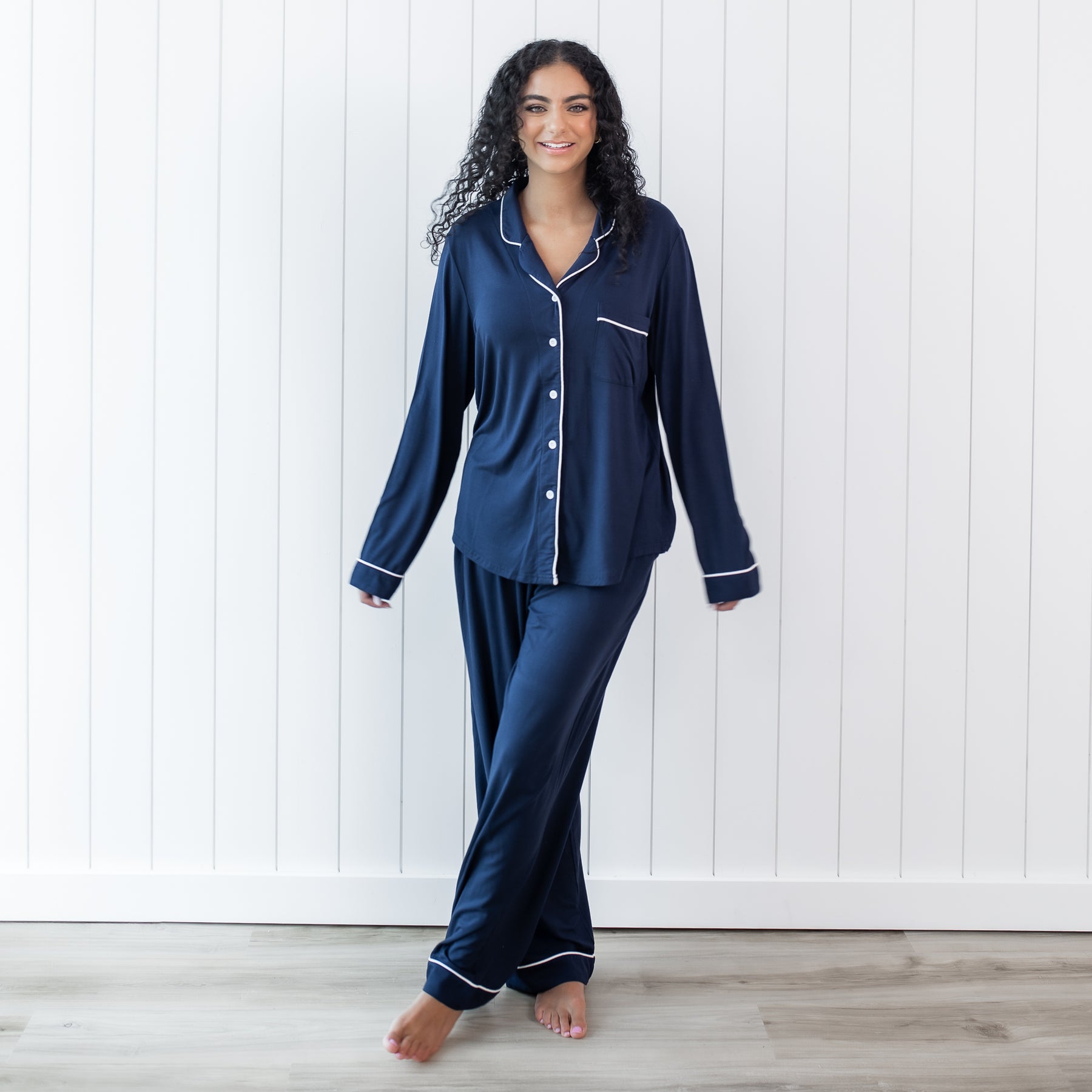 Female standing in front of a white paneled wall wearing the Long-Sleeved Women's Pajama Set in Navy with Cloud Trim