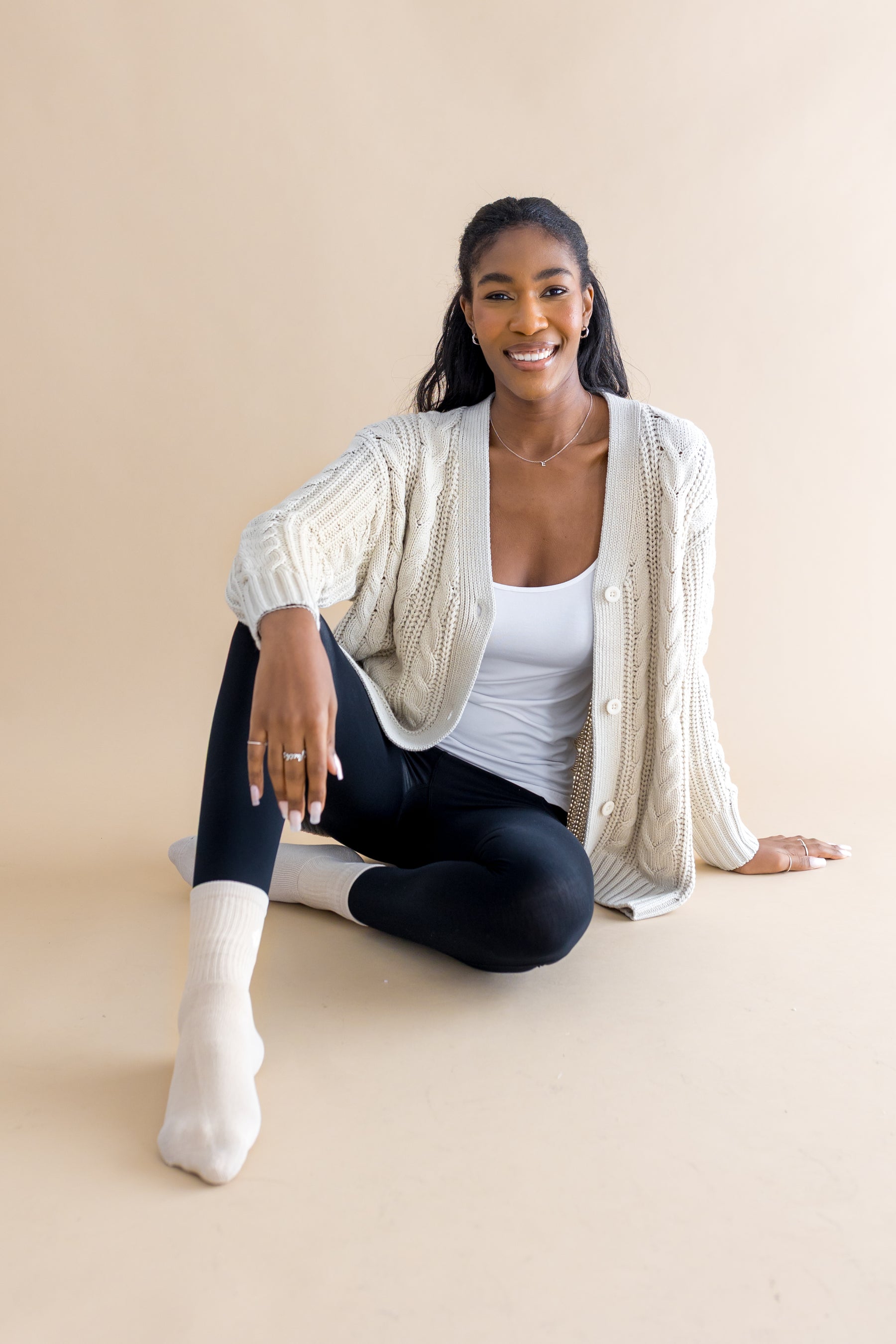 Smiling model sitting on the floor wearing the Adult Crew Socks in Oat with black tights, a white cami and cream colored cable knit cardigan