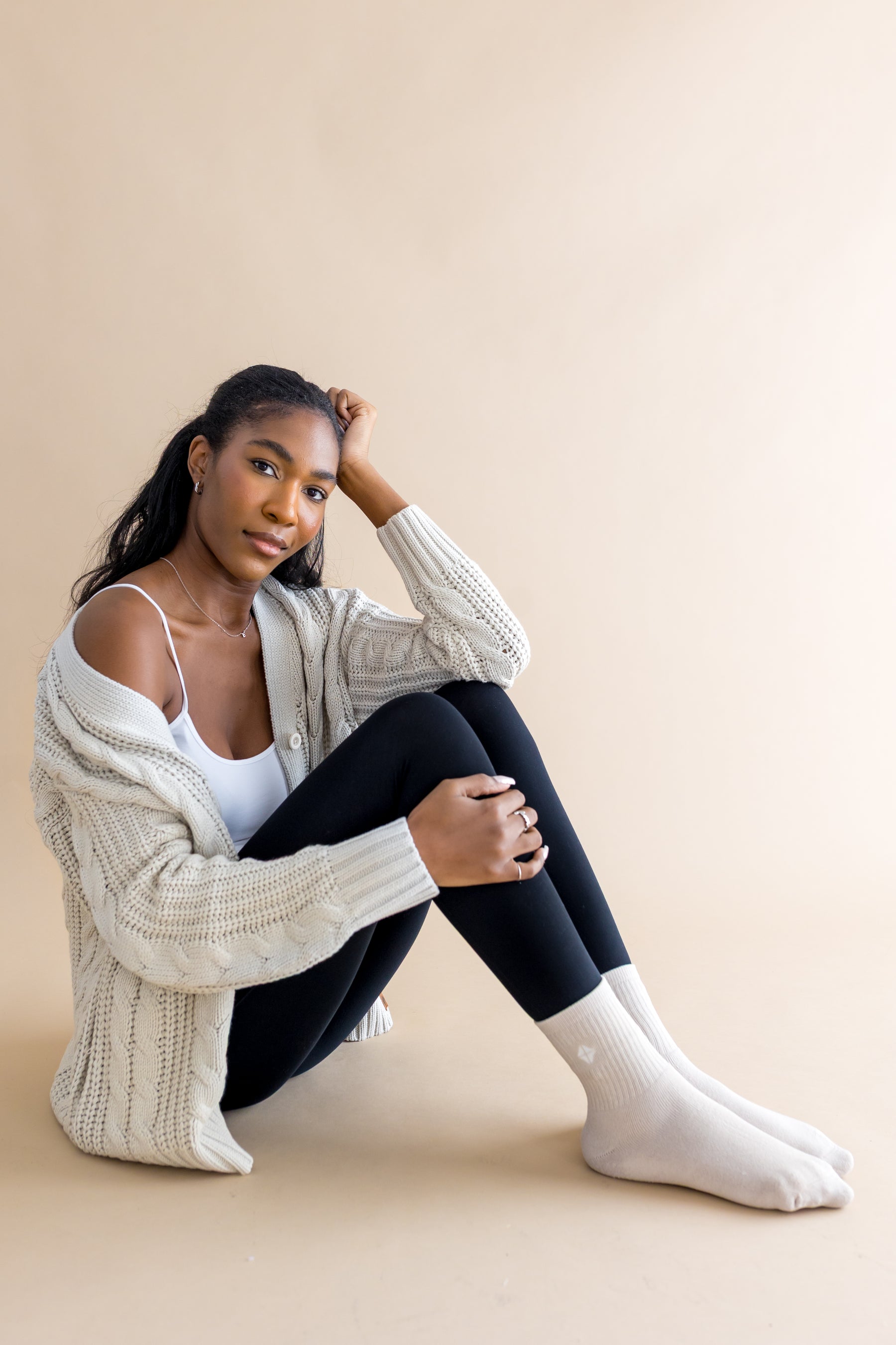 Female model sitting on the floor wearing the Adult Crew Socks in Oat with black leggings, a white cami and oat colored cable knit sweater