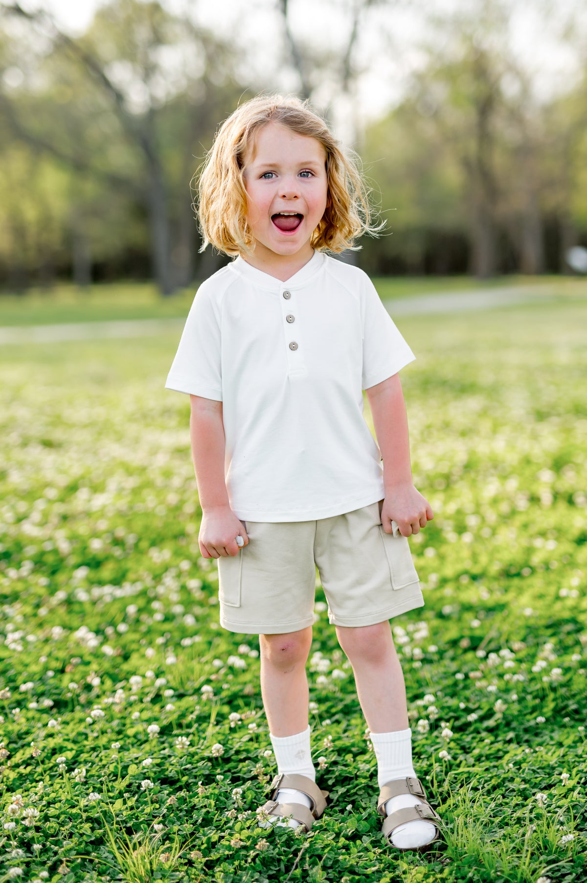 Young boy smiling standing in a field wearing the Cargo Short in Oat paired with a white short sleeve henley