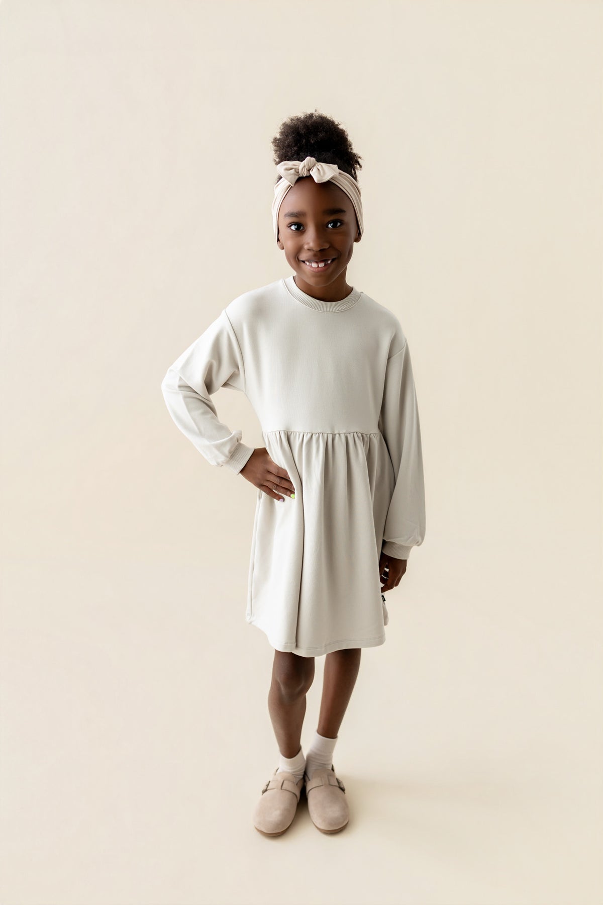 Girl wearing Long Sleeve Skater Dress in Oat color with knotted bow headband on a beige background