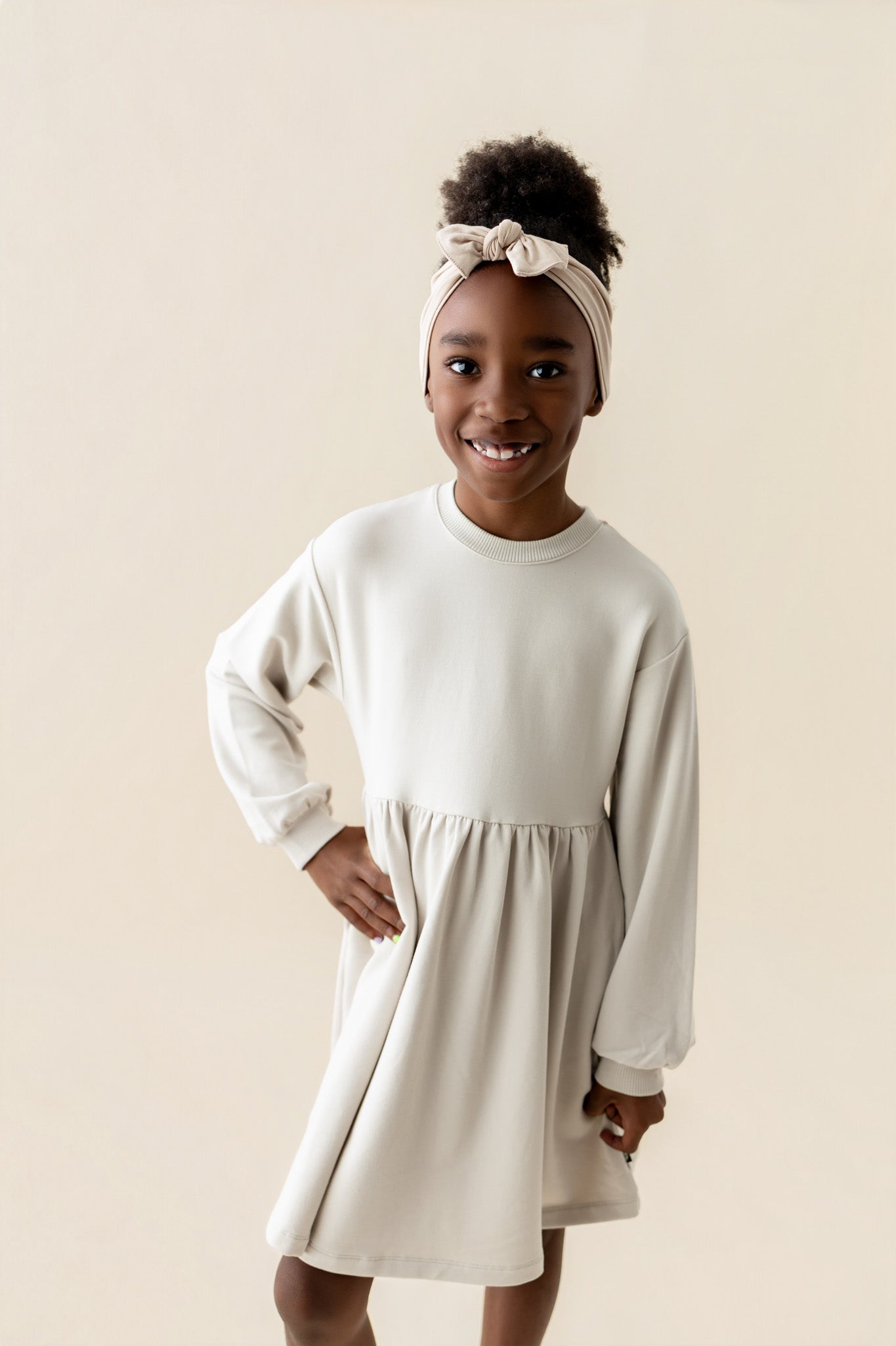 Girl wearing Long Sleeve Skater Dress in Oat color with knotted bow headband on a beige background