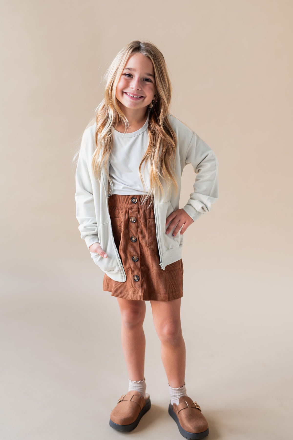 Girl smiling in Zip-Up Hoodie in Oat color over a rust color skirt and white T-shirt on a white background
