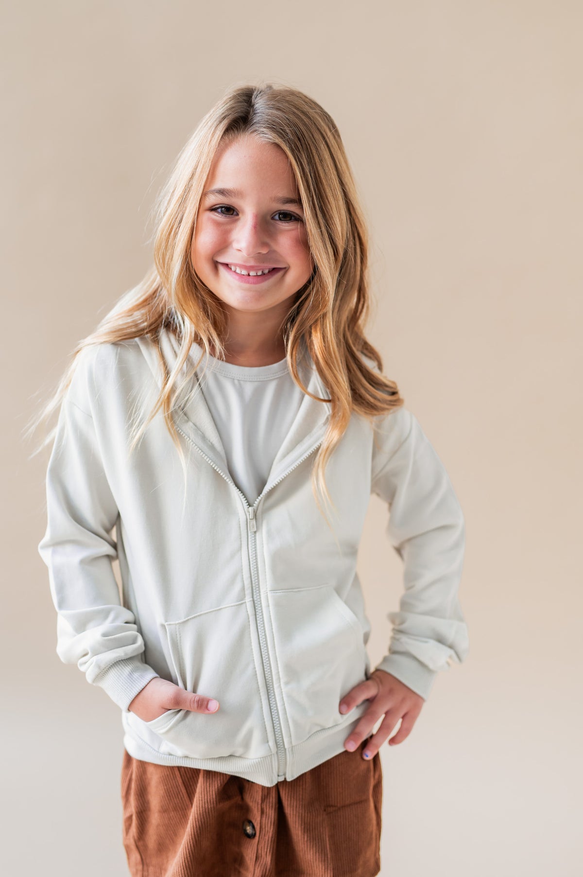 Girl posing in Zip-Up Hoodie in Oat color over a rust color skirt and white T-shirt on a white background