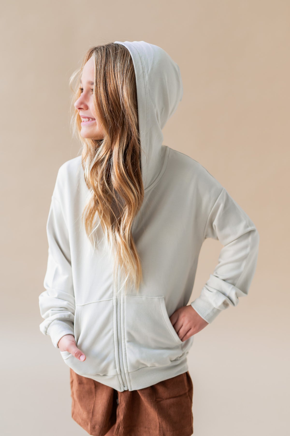 Girl wearing Zip-Up Hoodie in Oat color over a rust color skirt on a white background