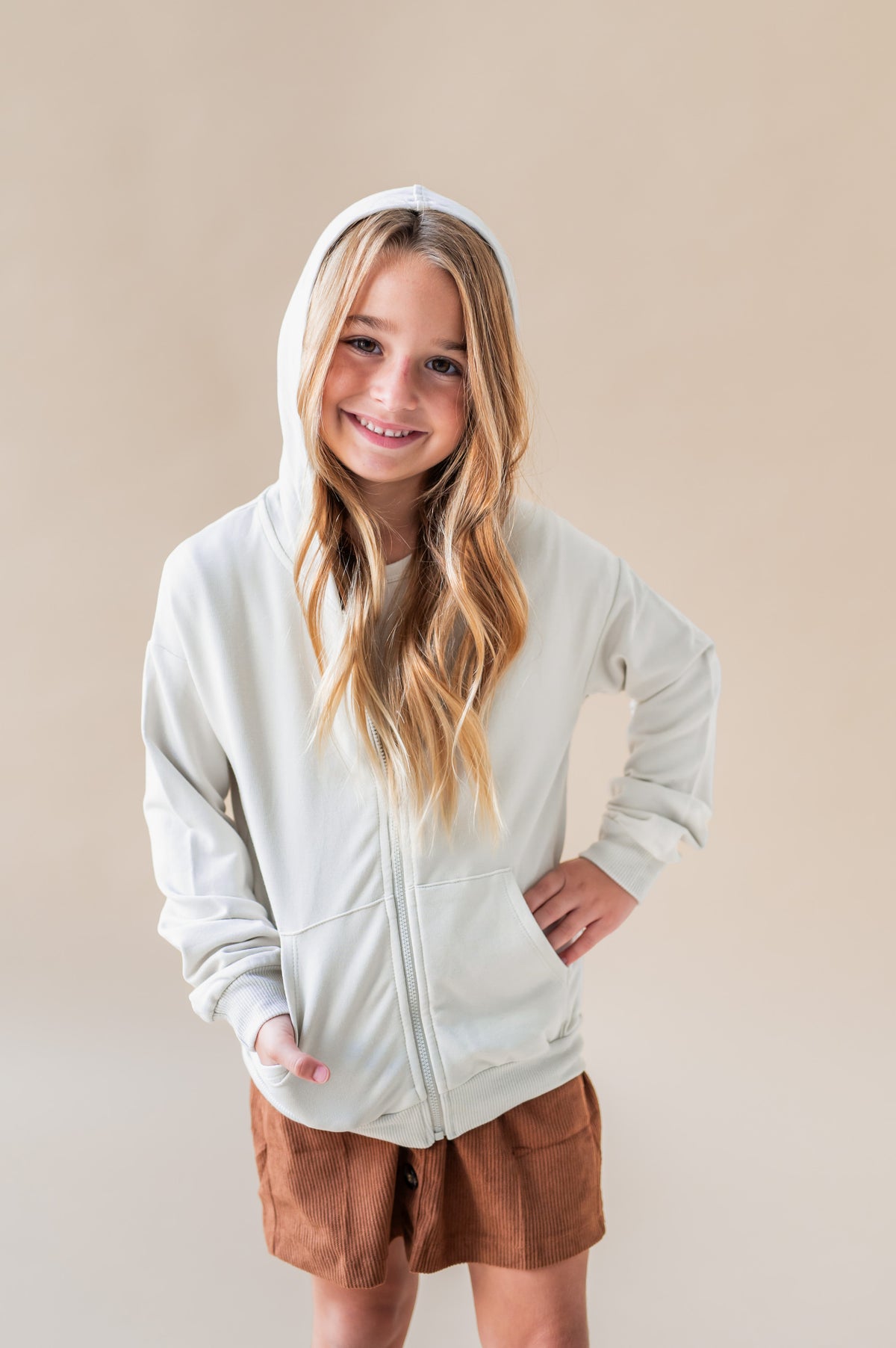 Girl wearing Zip-Up Hoodie in Oat color over a rust color skirt on a white background
