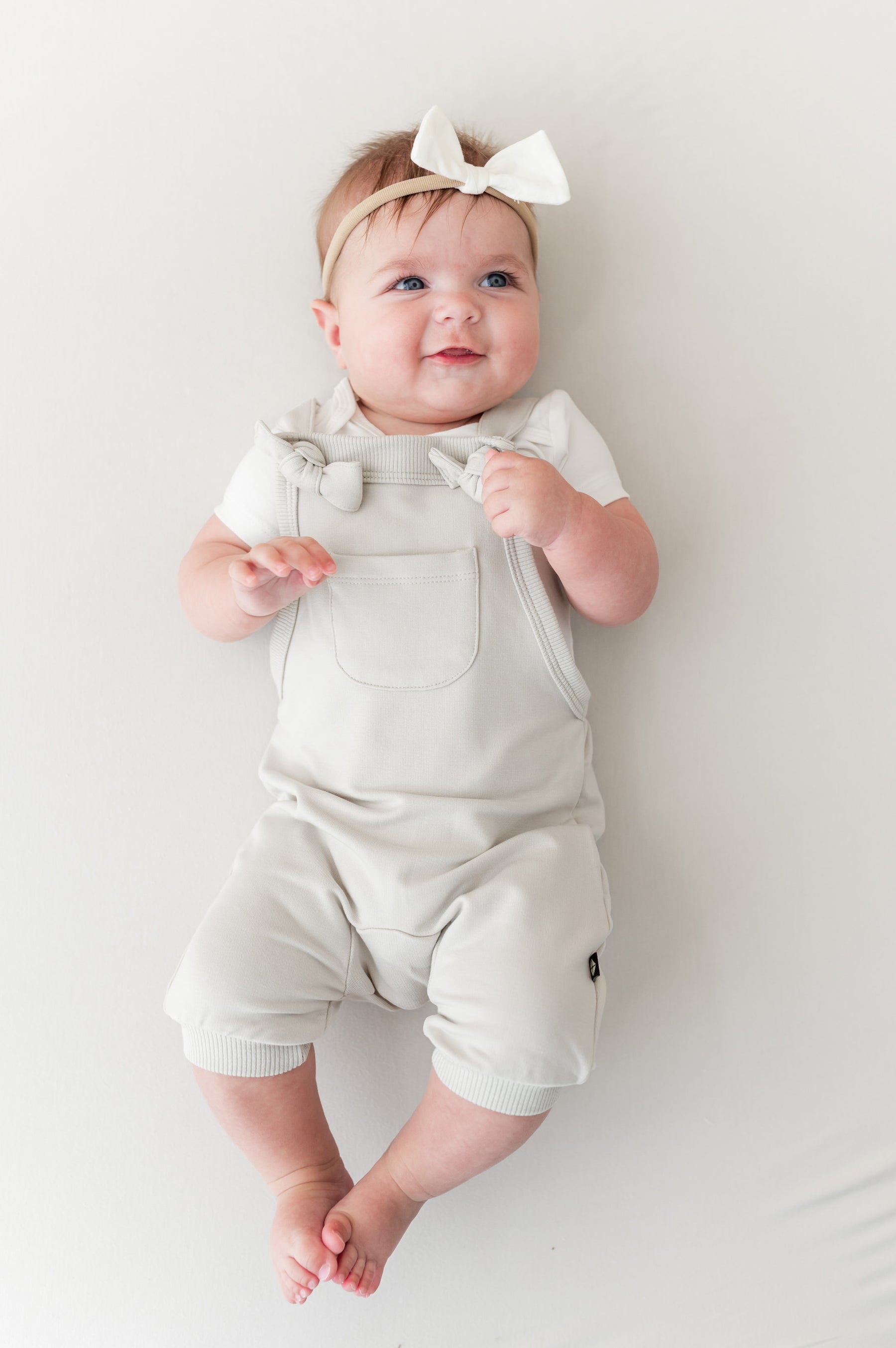 Baby wearing Overall in Oat color with white bow headband on a beige background