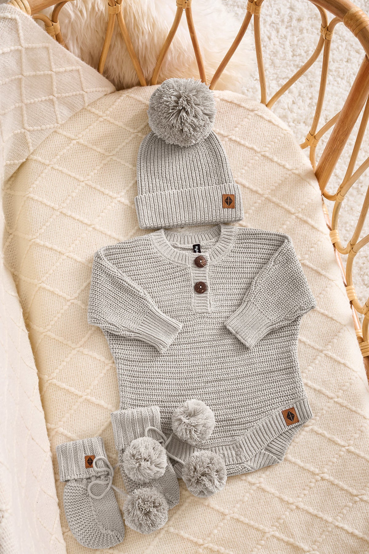A baby knit outfit set in a wicker crib, featuring a light gray long-sleeve Henley bodysuit with three brown buttons, a matching gray knit beanie with a pom-pom, and gray booties with pom-poms.