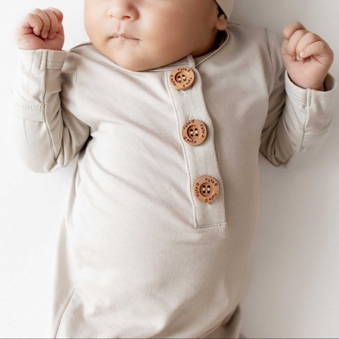 Close up of Baby in oat colored knotted gown outfit that has wooden buttons and a matching hat