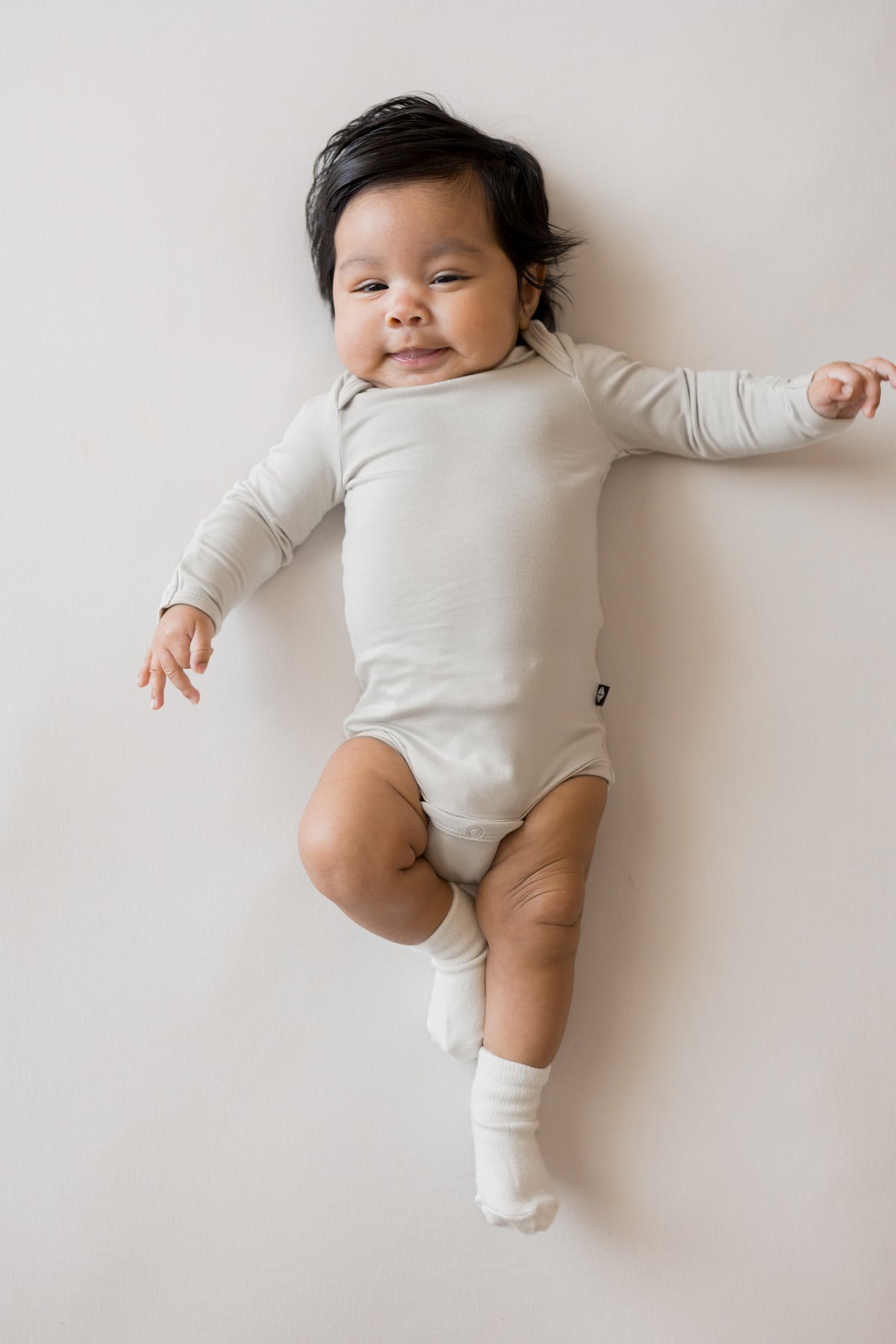 Baby wearing Long Sleeve Bodysuit in Oat color and white socks on a beige background