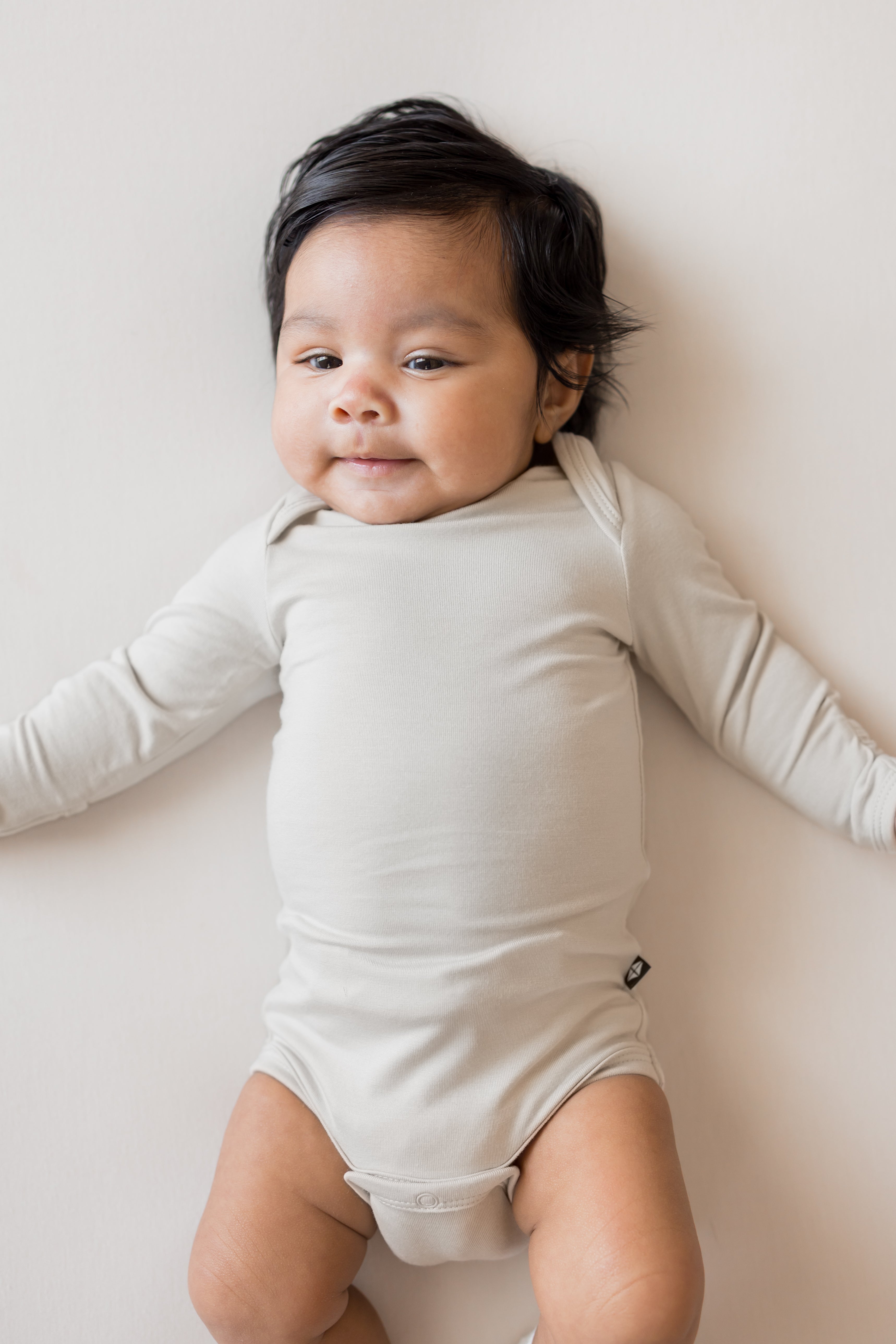 Baby wearing Long Sleeve Bodysuit in Oat color lying on a beige background