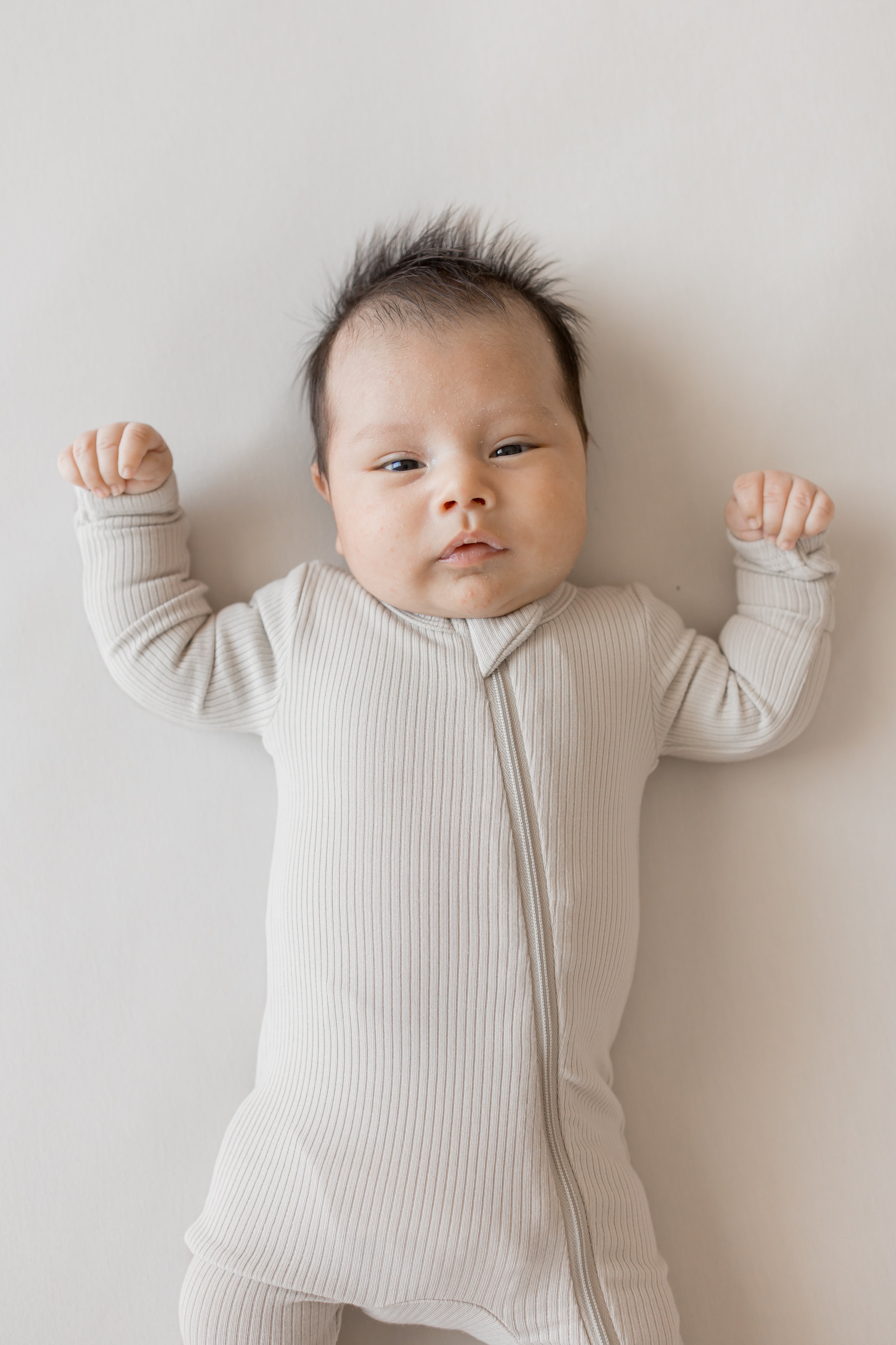 Close up of Baby wearing Ribbed Zipper Romper in Oat color on a beige background