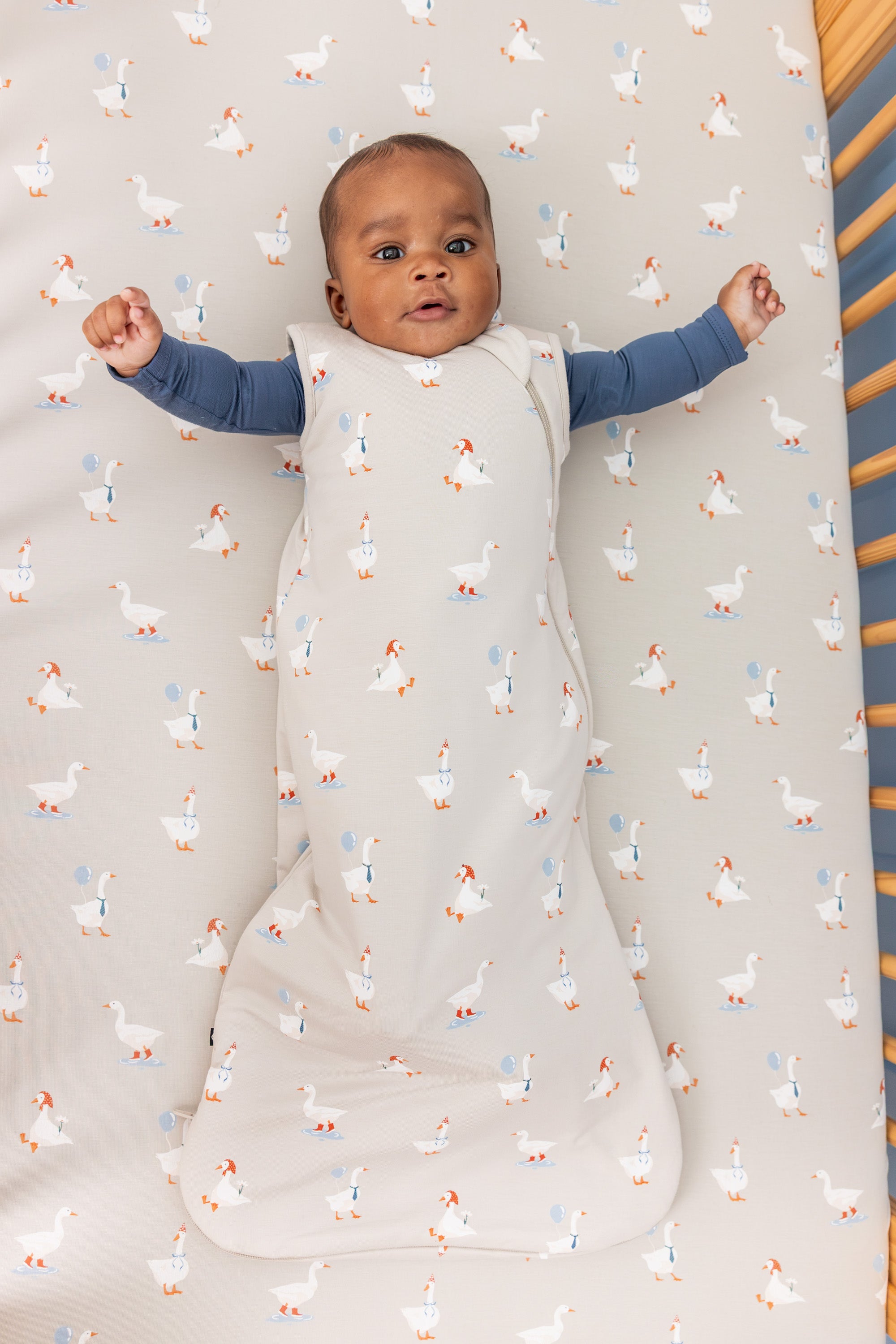 Baby wearing Sleep Bag in Oat Silly Goose 1.0 print over oxford zippered romper lying in a crib over oat silly goose print crib sheet